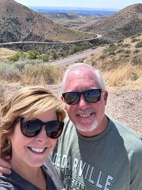A smiling couple taking a selfie outdoors in a hilly area with dry grass and a winding road in the background, wearing sunglasses and casual clothing.