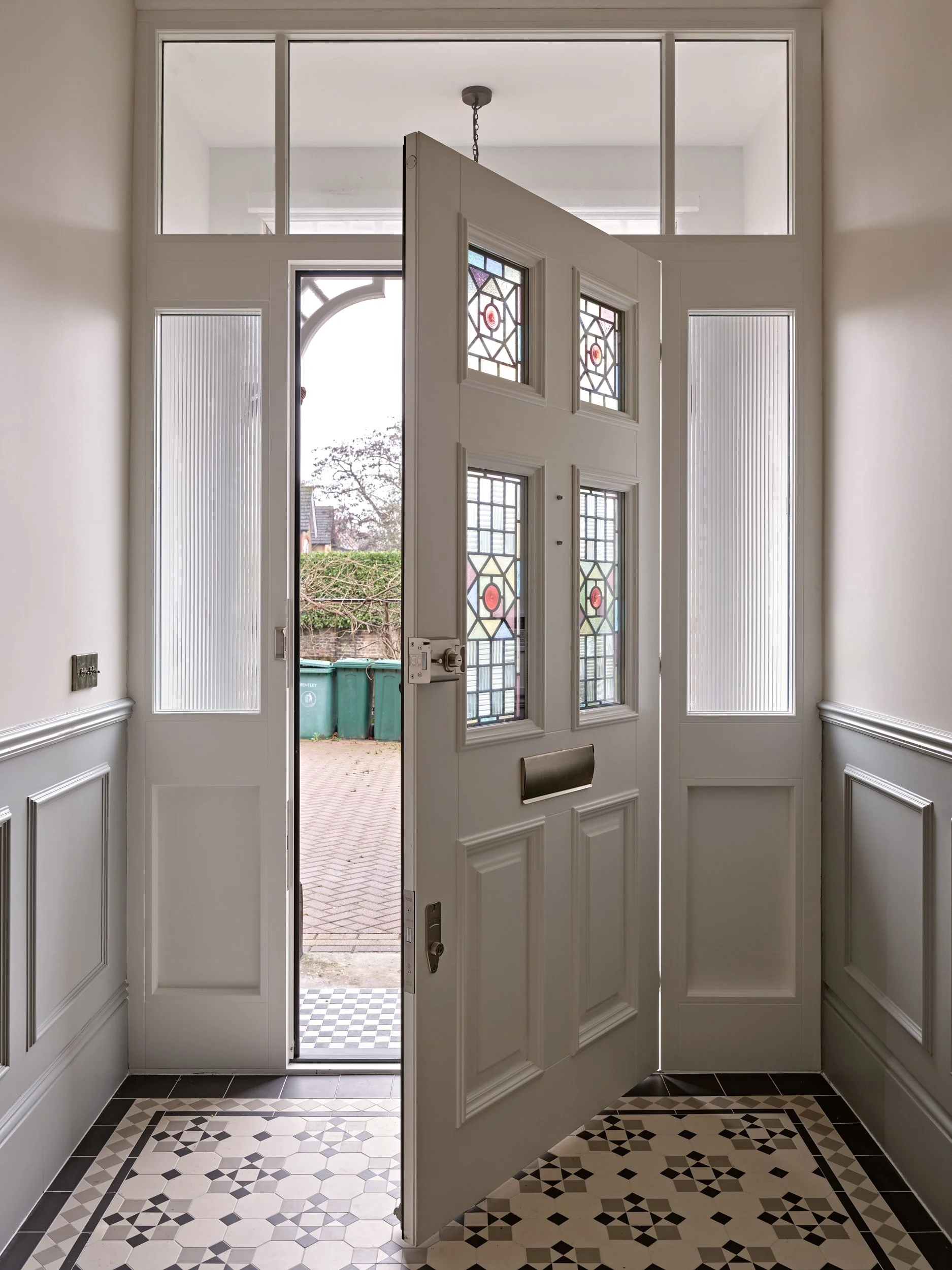 View of a front door with stained glass window panels, open to reveal an outside brick patio with three green trash bins, surrounded by hedge and trees.