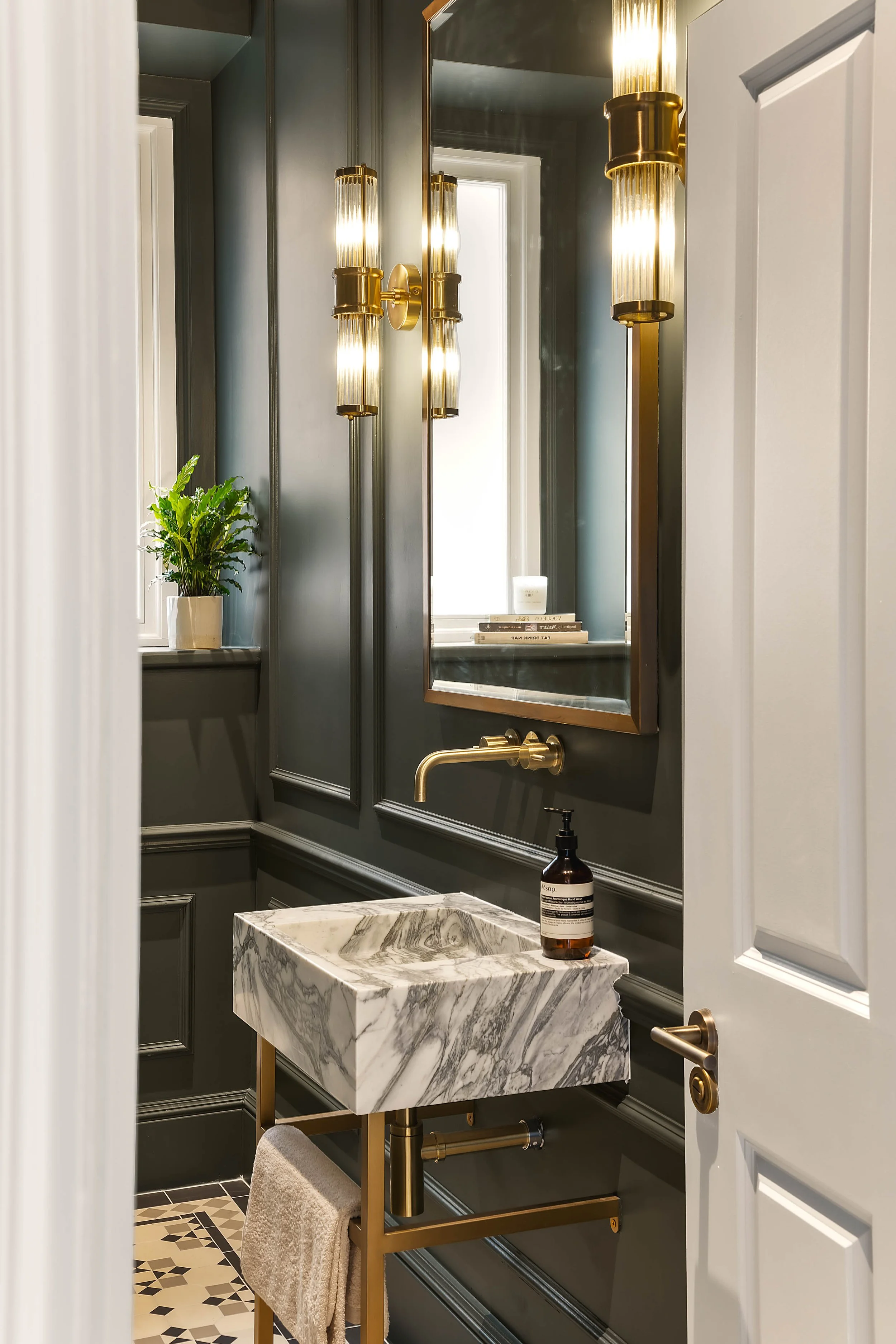 A small bathroom with dark gray paneled walls and a marble sink. There is a gold faucet, a large mirror, and decorative gold wall sconces. A potted plant is on the windowsill, and a towel hangs on a gold towel rack below the sink.