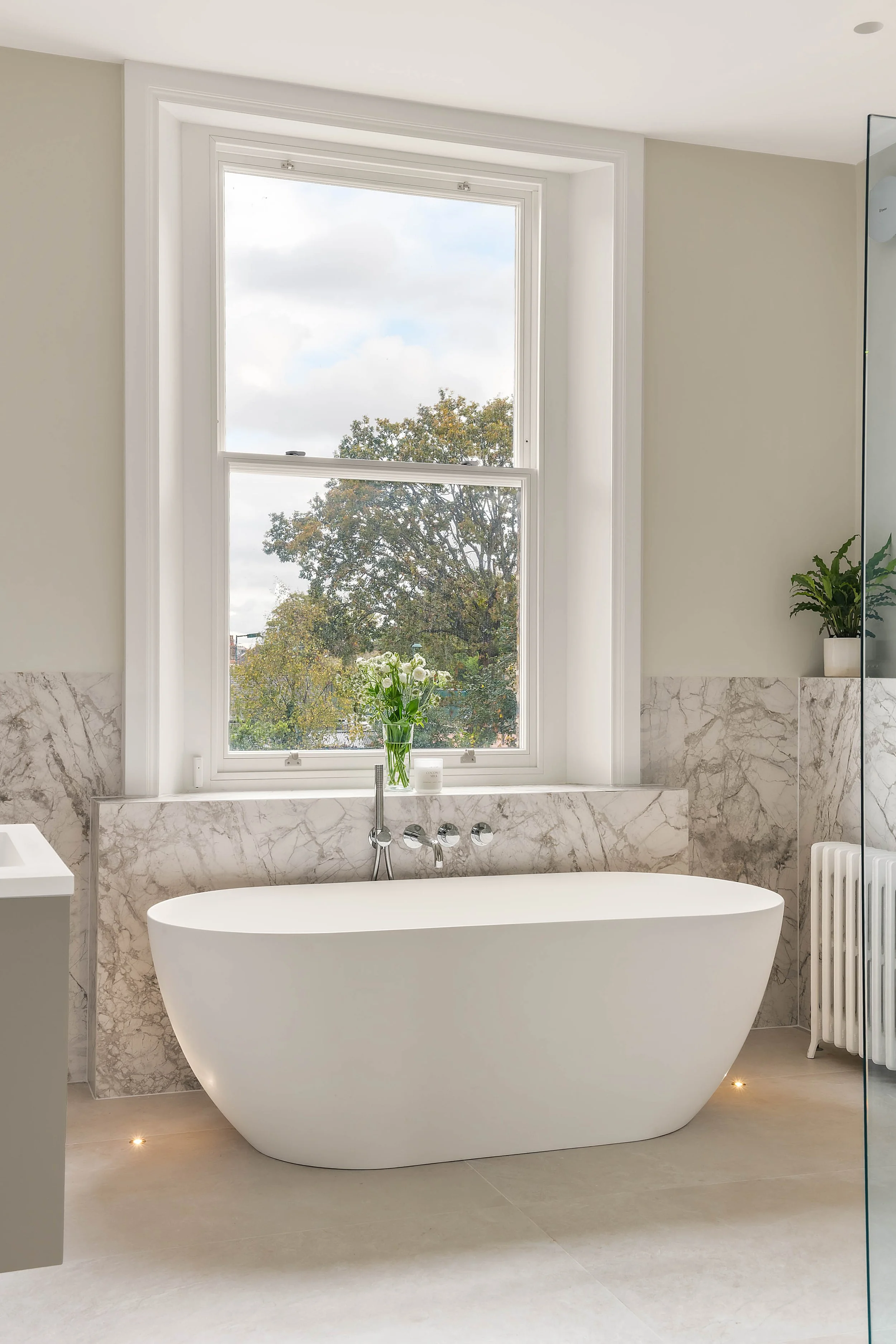 Modern bathroom with a freestanding bathtub beneath a large window, decorated with a small plant and flowers, featuring marble accents and neutral tones.