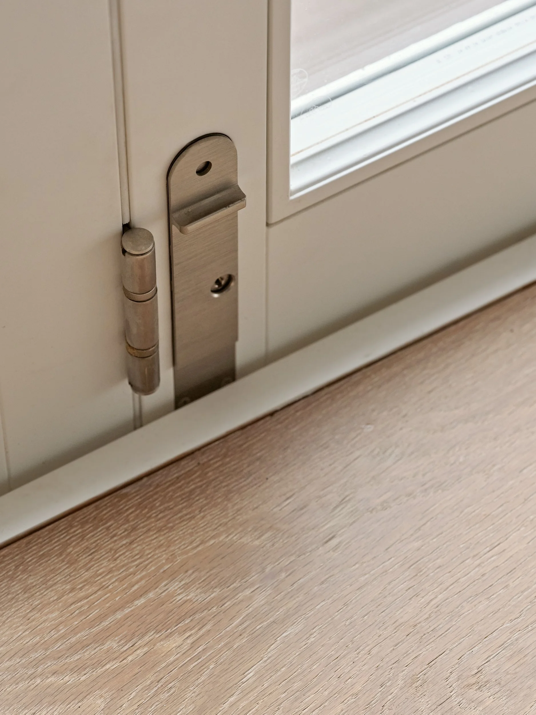 Close-up of a door hinge and latch mechanism on a white door next to a window with a wooden floor beneath.