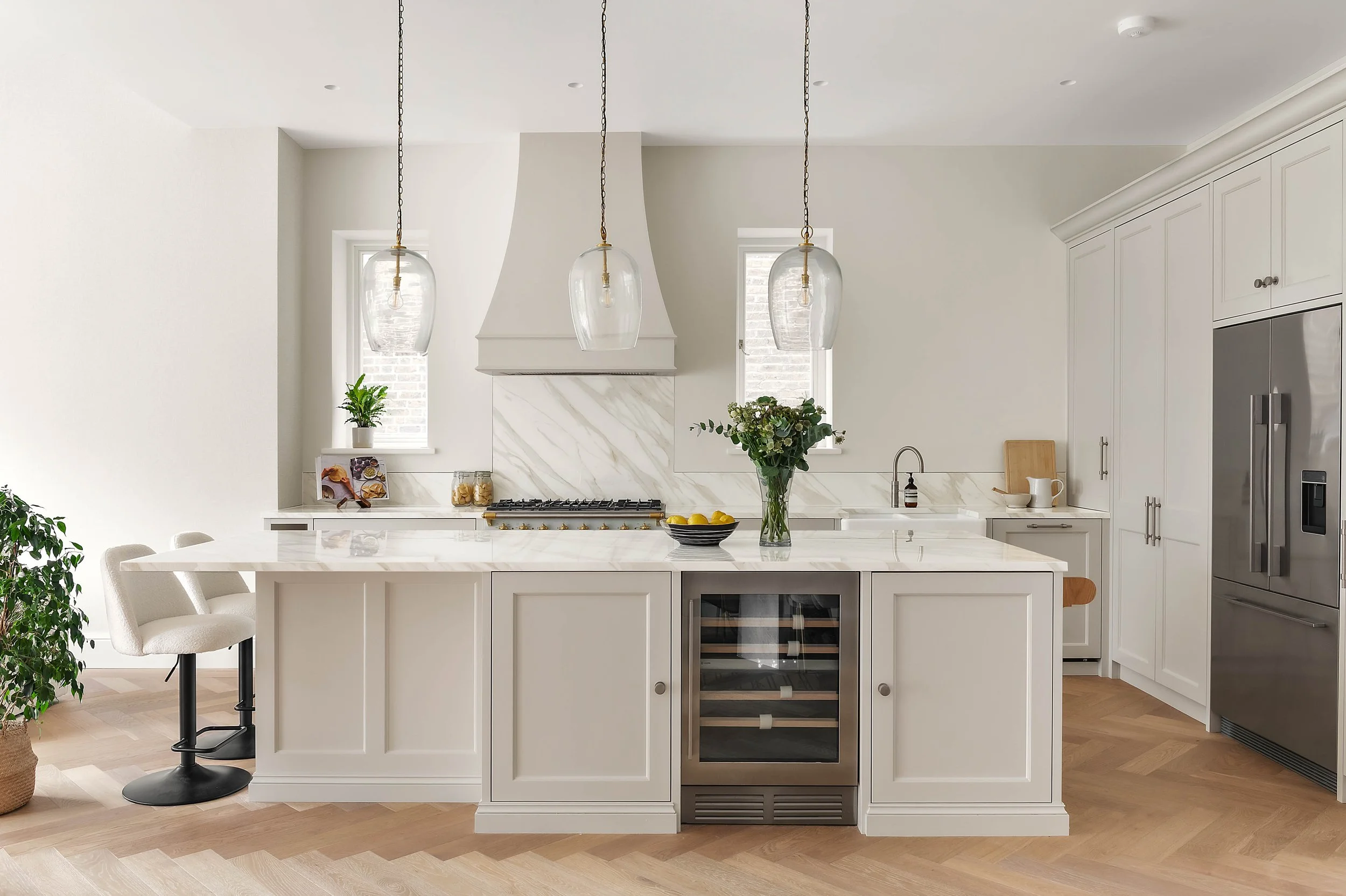 Modern white kitchen with marble island, hanging pendant lights, a wine cooler, and a vase of flowers.