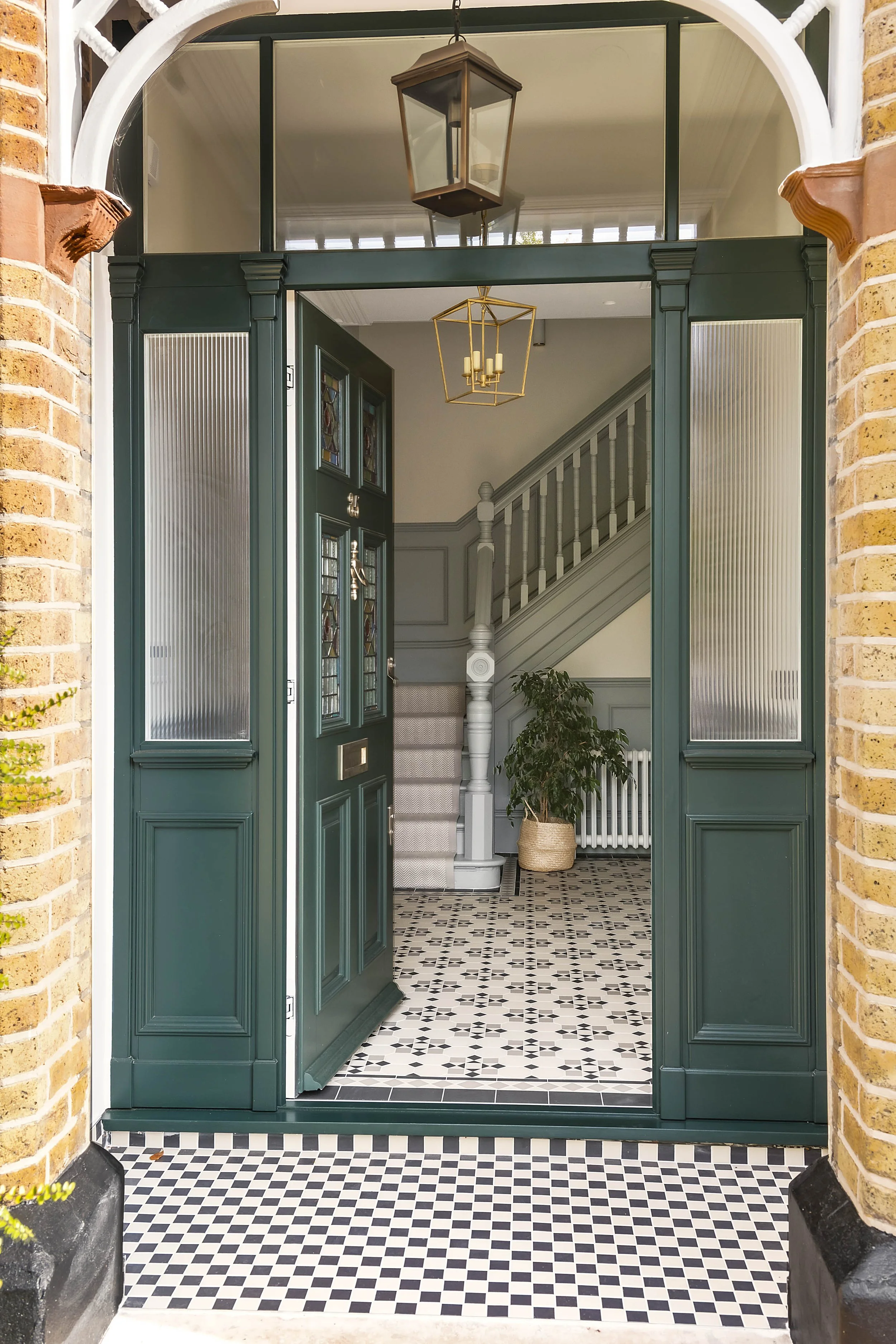 The front door of a house with dark green paint and stained glass panels. The door opens to a foyer with patterned black-and-white tile floor, a staircase with a white banister, a potted plant, and contemporary light fixtures hanging from the ceiling