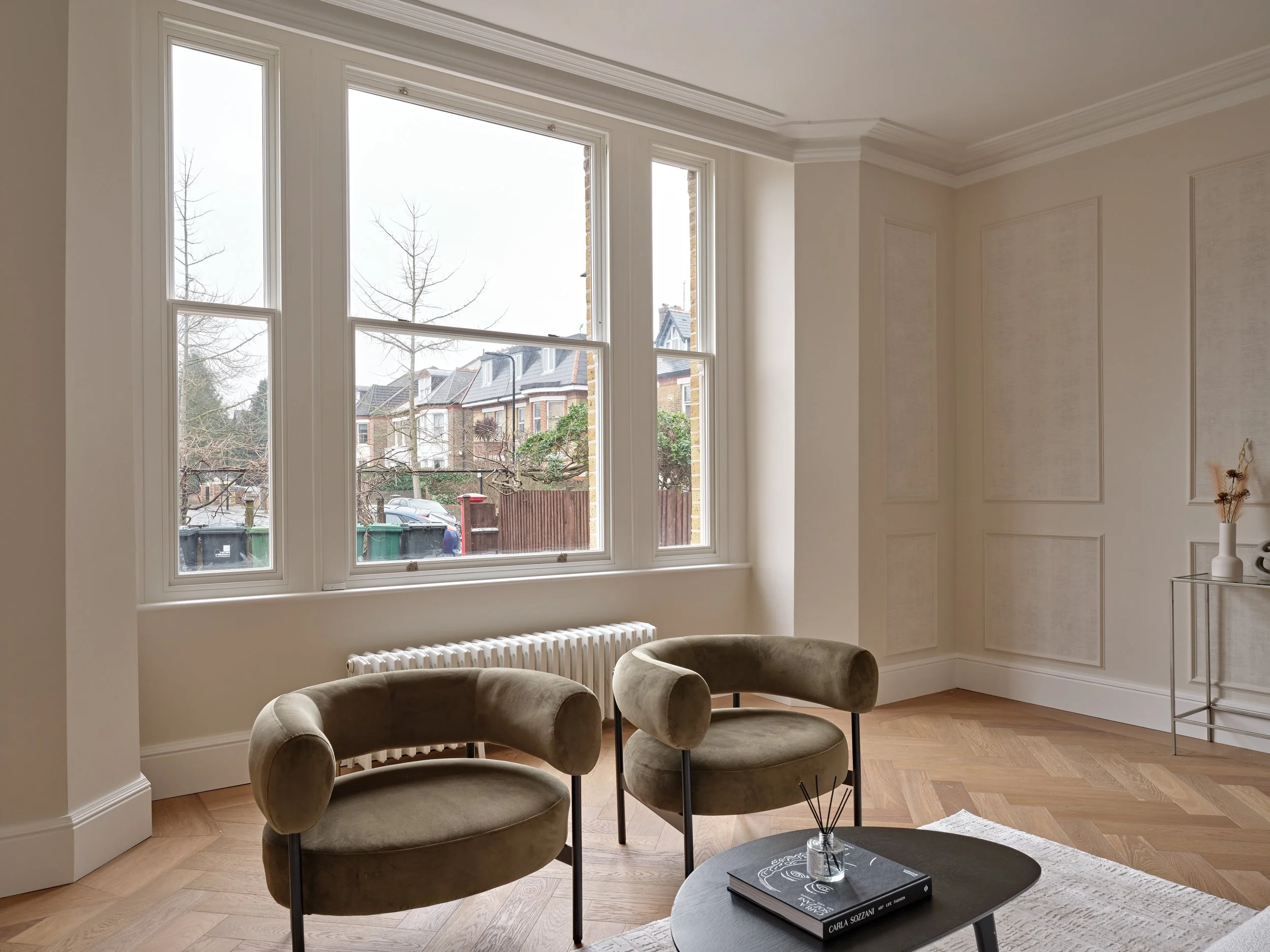 A bright living room with two modern beige velvet armchairs, a black coffee table with a small decorative item and books, large windows revealing a street with houses, and light-colored walls with decorative paneling.