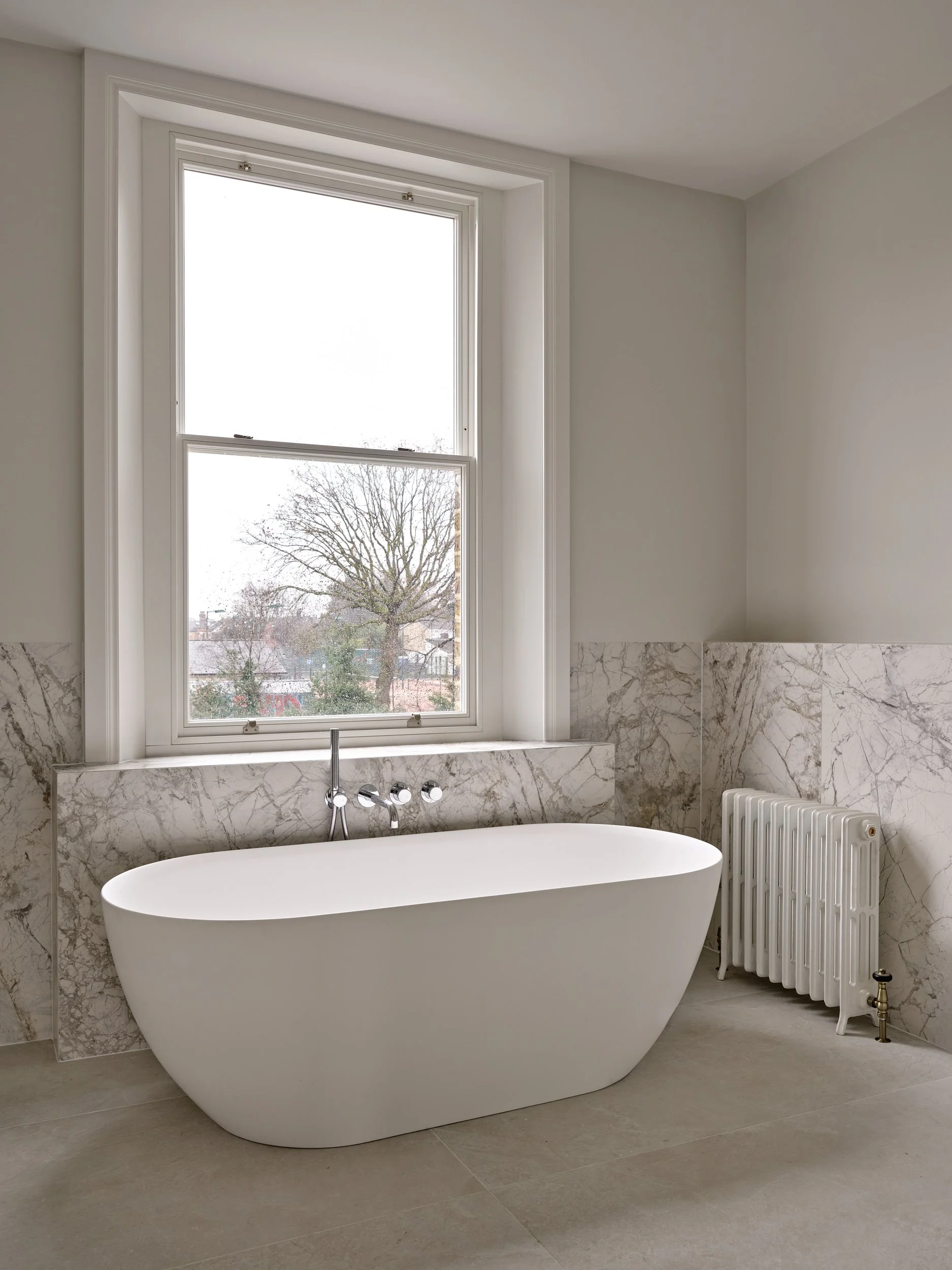 Modern bathtub in a bright bathroom with large window, marble backsplash, and radiator.
