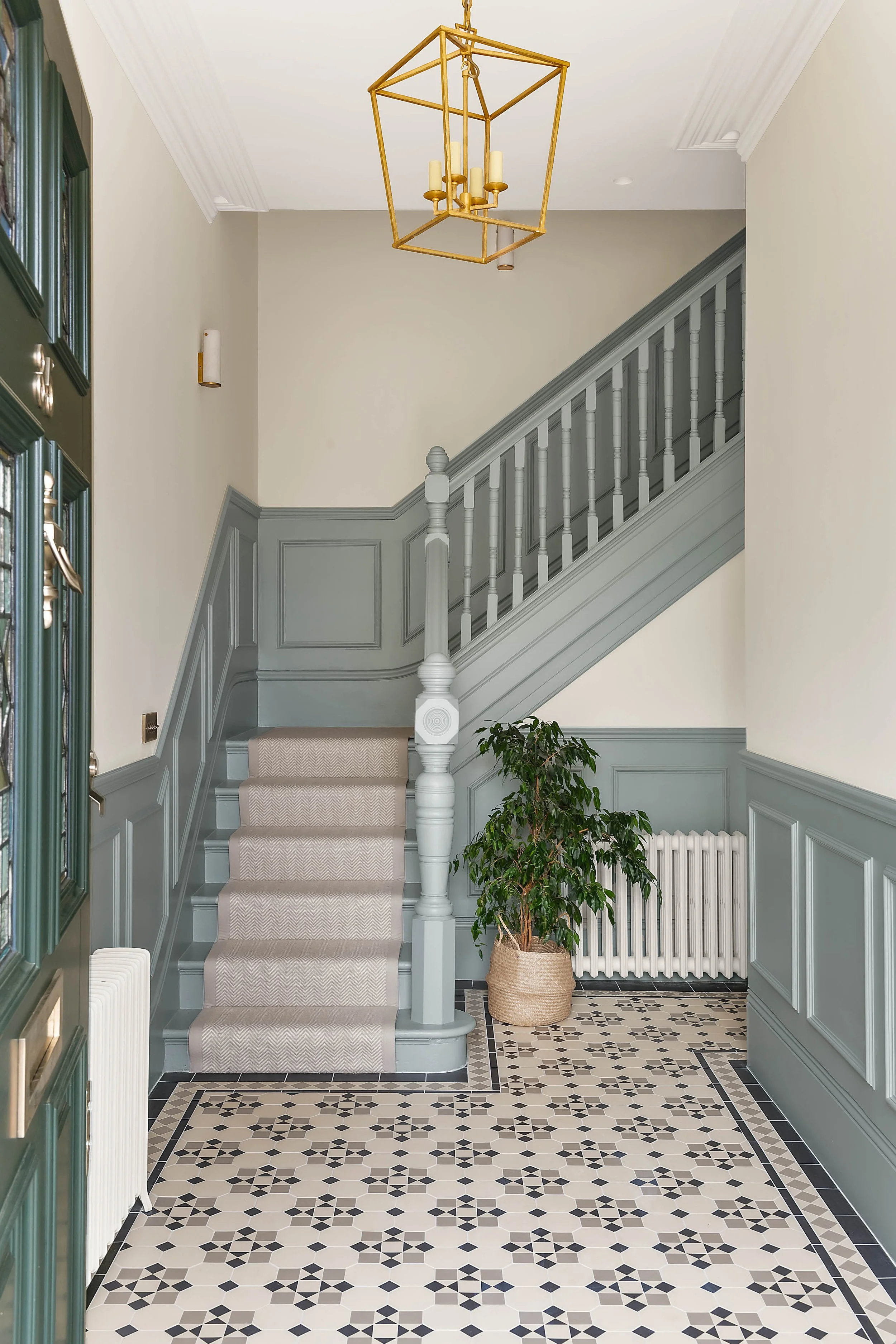 Entryway with patterned tile floor, staircase with gray woodwork and beige carpet, a potted plant, radiator, and a gold geometric chandelier hanging from the ceiling.