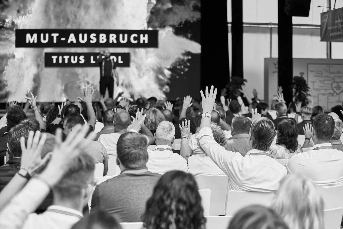 Eine große Menschenmenge bei einer Konferenz mit vielen Personen, die ihre Hände heben, vor einer großen Leinwand, auf der der Text 'Mut-Ausbruch' und 'Titus Lindl' zu sehen ist.