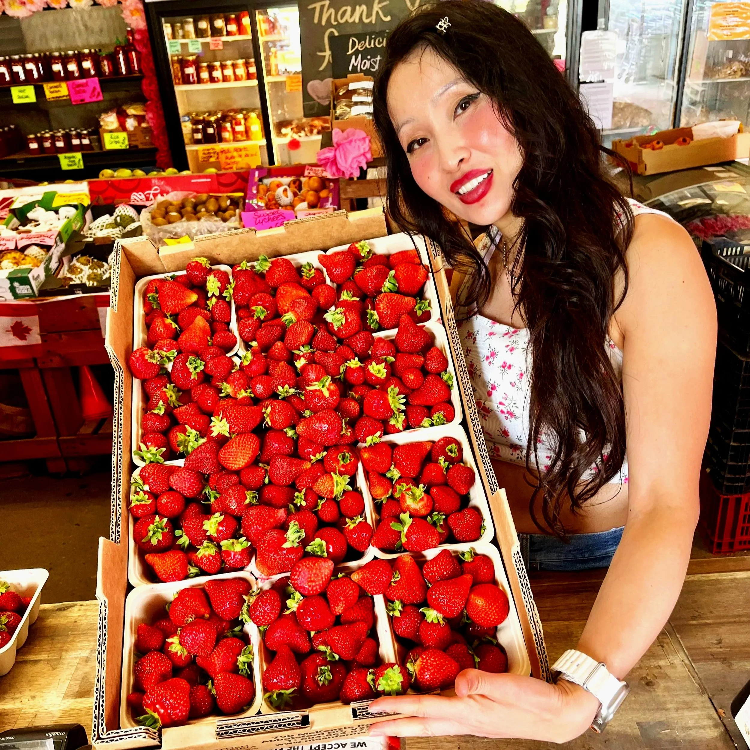 A woman holding a basket of fresh strawberries at a grocery store or market. The background displays shelves with jars and signs, and there are other fruit boxes around.
