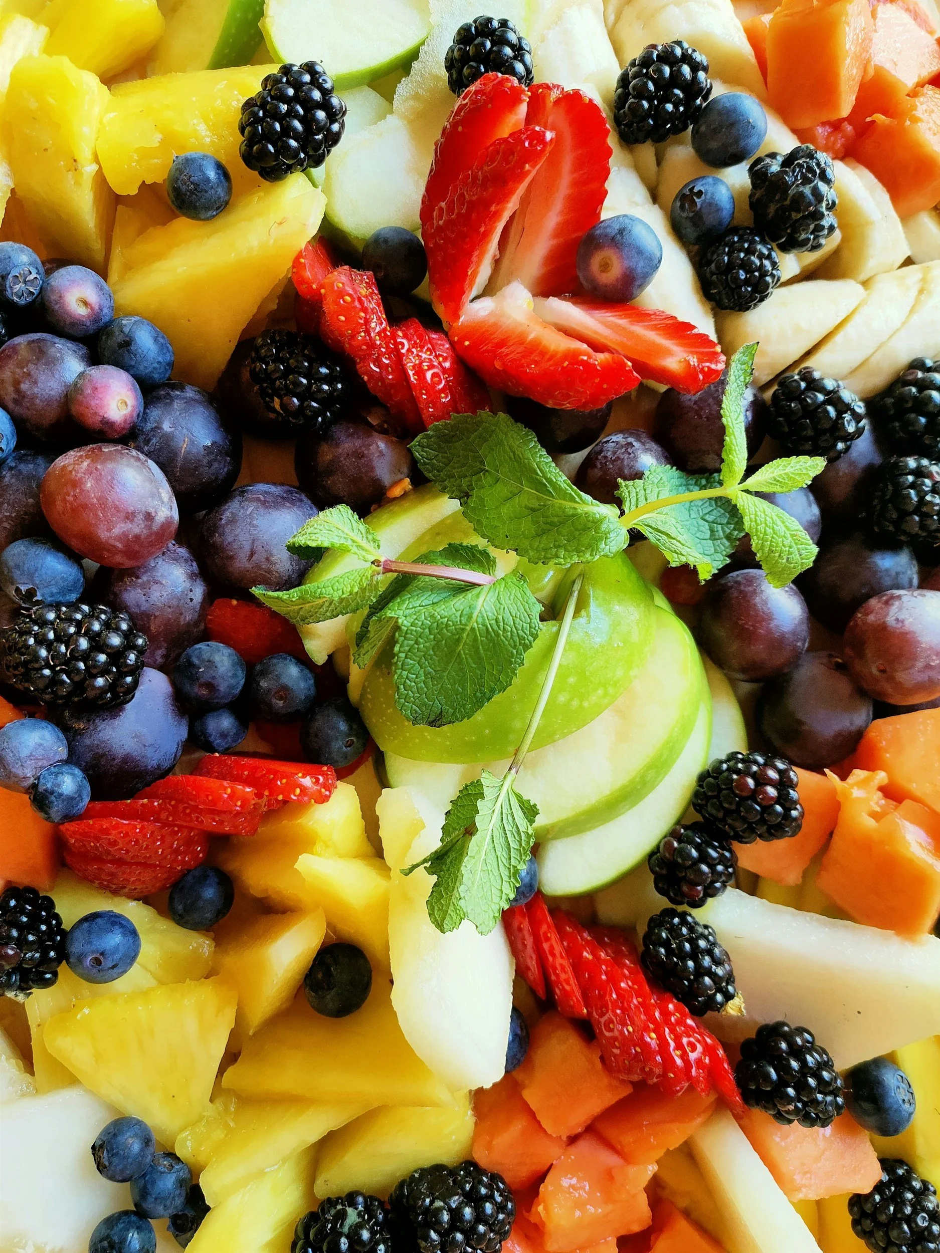 A colorful arrangement of sliced fruits including pineapple, blueberries, strawberries, blackberries, green apple slices, cantaloupe, and honeydew, garnished with fresh mint leaves.