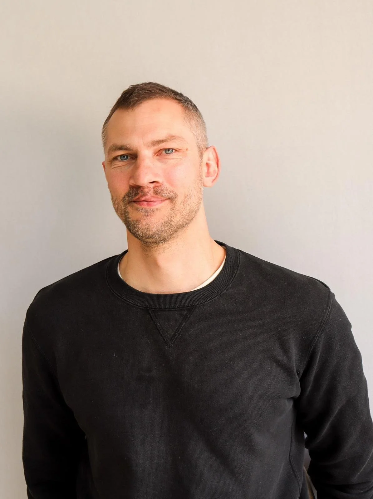 A man with short brown hair, blue eyes, and a beard, wearing a black sweatshirt, standing against a plain light-colored wall.