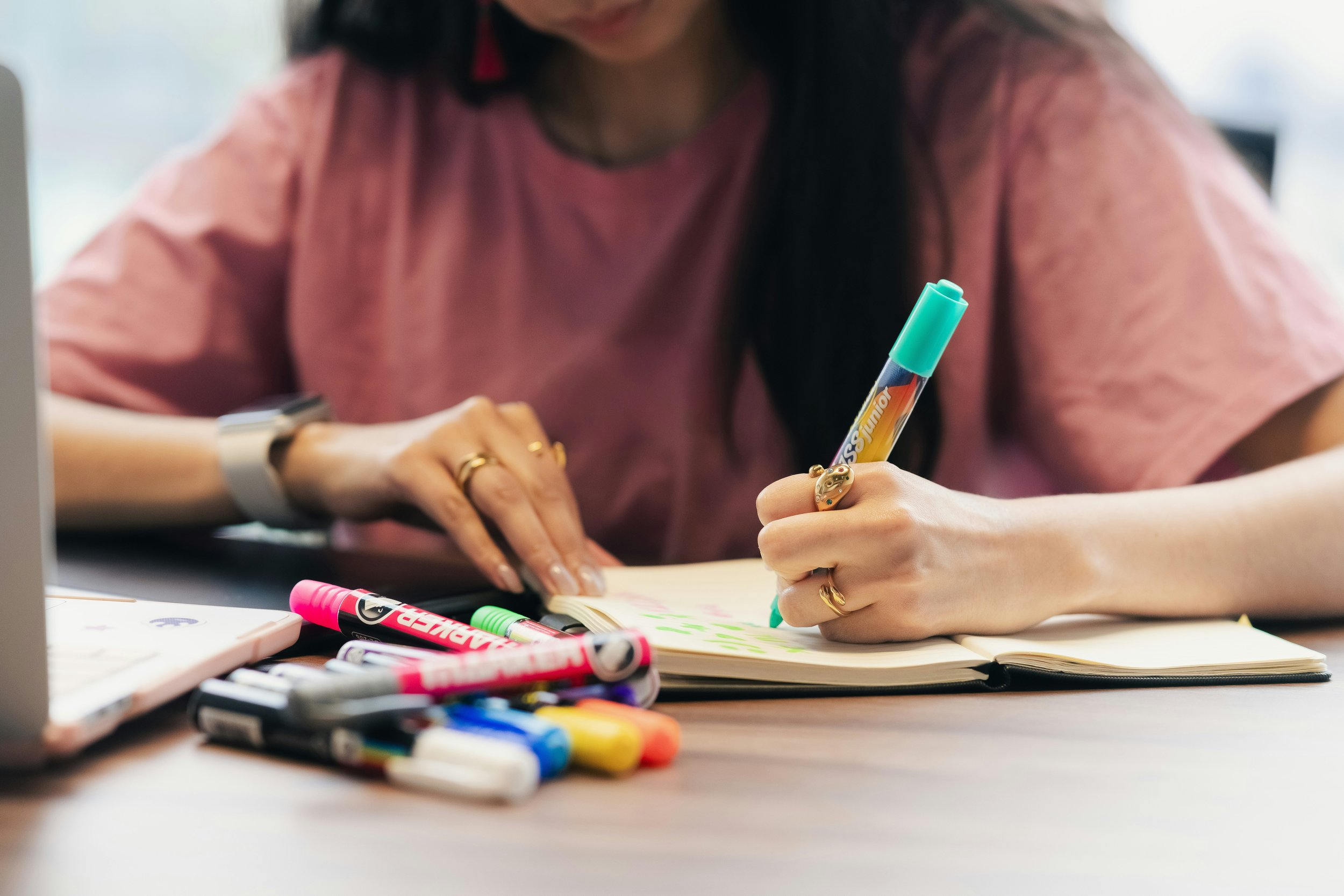 Person wearing a pink shirt writing in a notebook with colorful markers on a table.