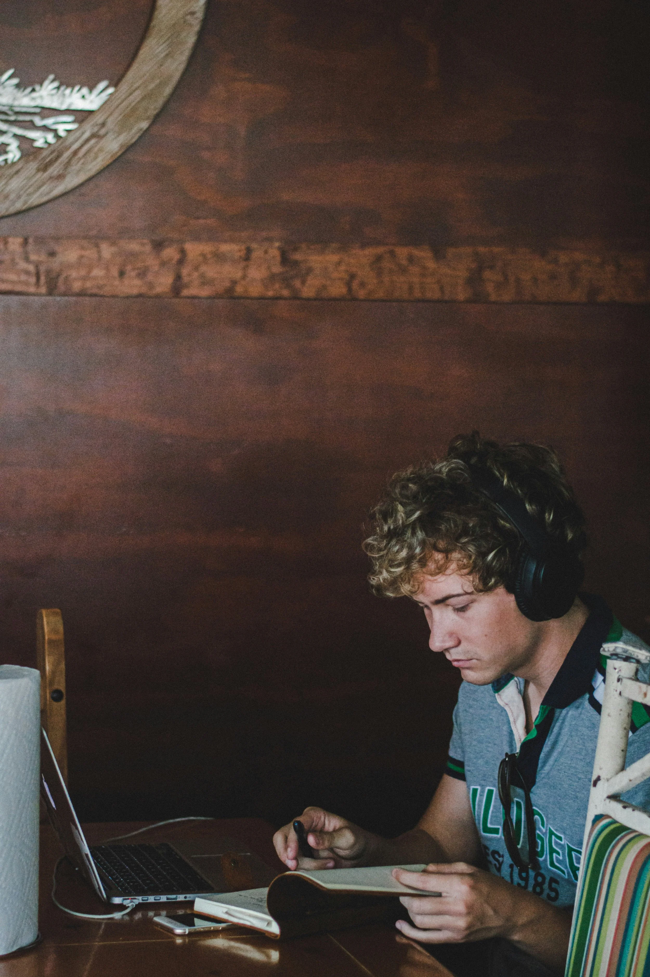A young man with curly hair wearing headphones, a graphic T-shirt, and glasses hanging from his shirt collar, sitting at a wooden table working on a laptop, reading a book, and holding a smartphone in his hand in a cozy room with a wooden ceiling.