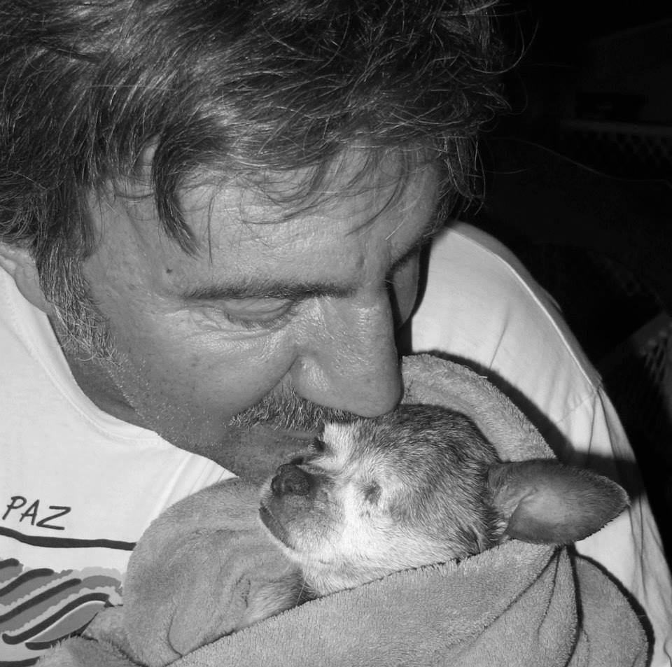 A man with gray hair and a beard holding and kissing a small sleeping puppy wrapped in a blanket.