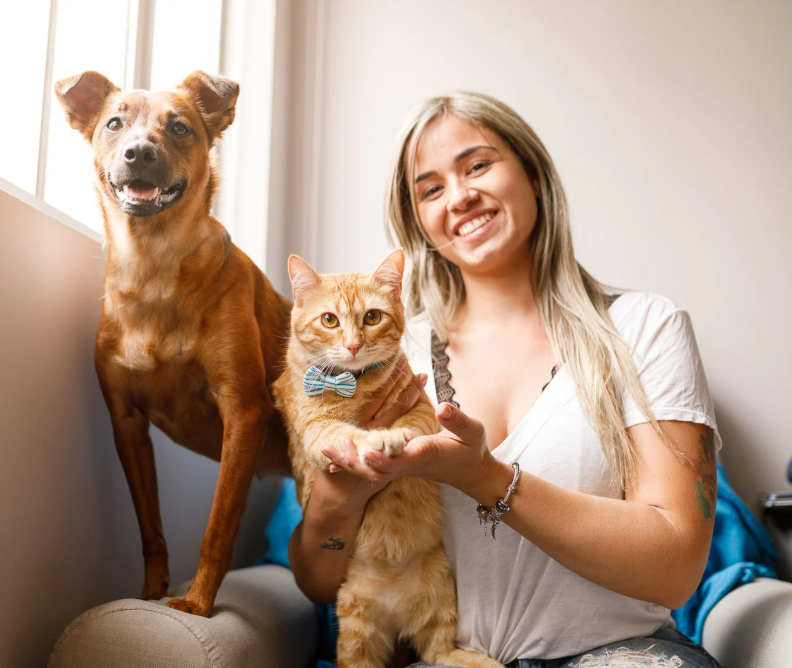 A smiling woman holding an orange tabby cat with a bow tie, and a brown dog standing on a sofa next to her in a bright room.