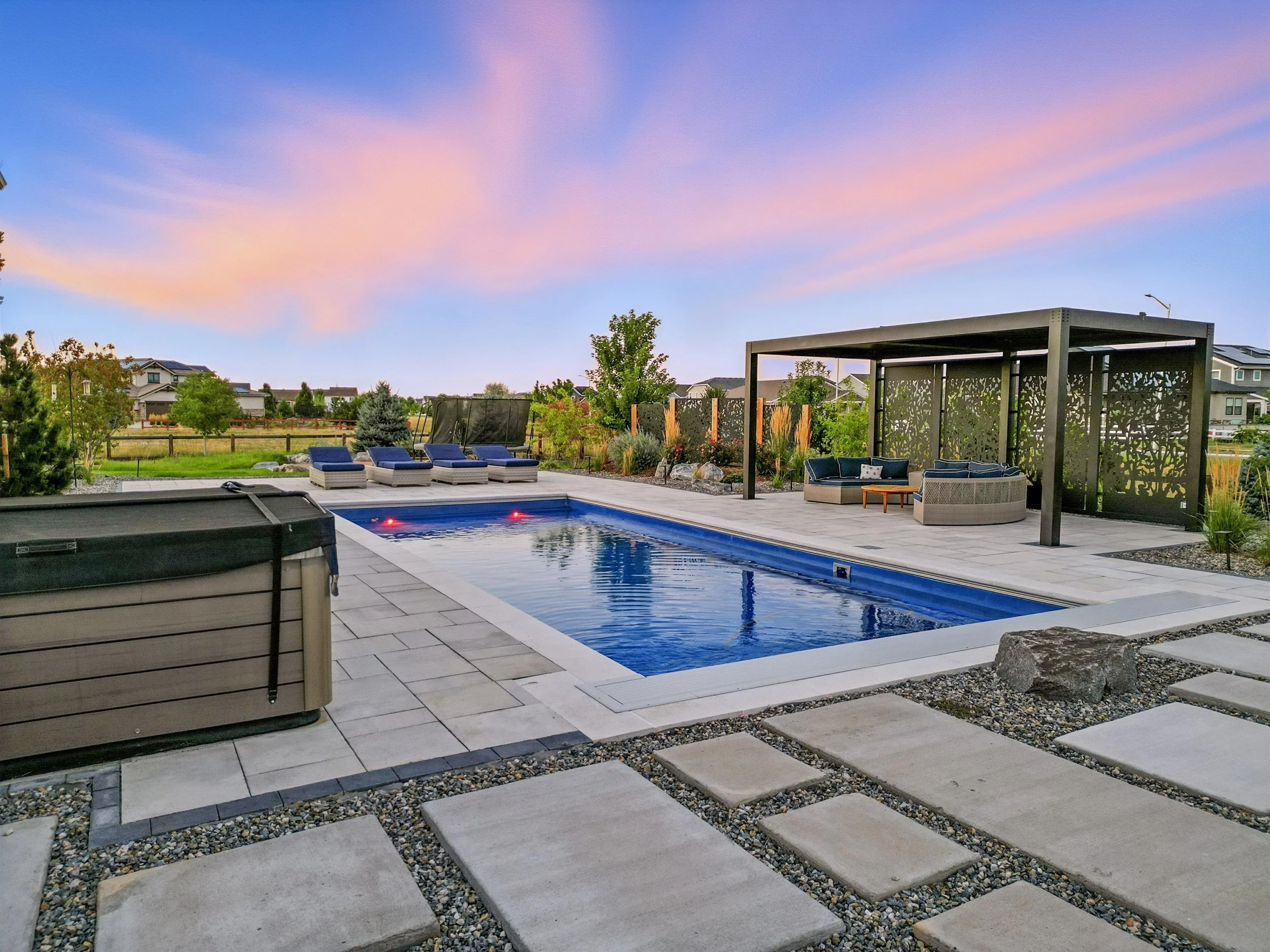 A backyard swimming pool area with lounge chairs, a hot tub, and a shaded seating area at dusk, with a colorful sky in the background.