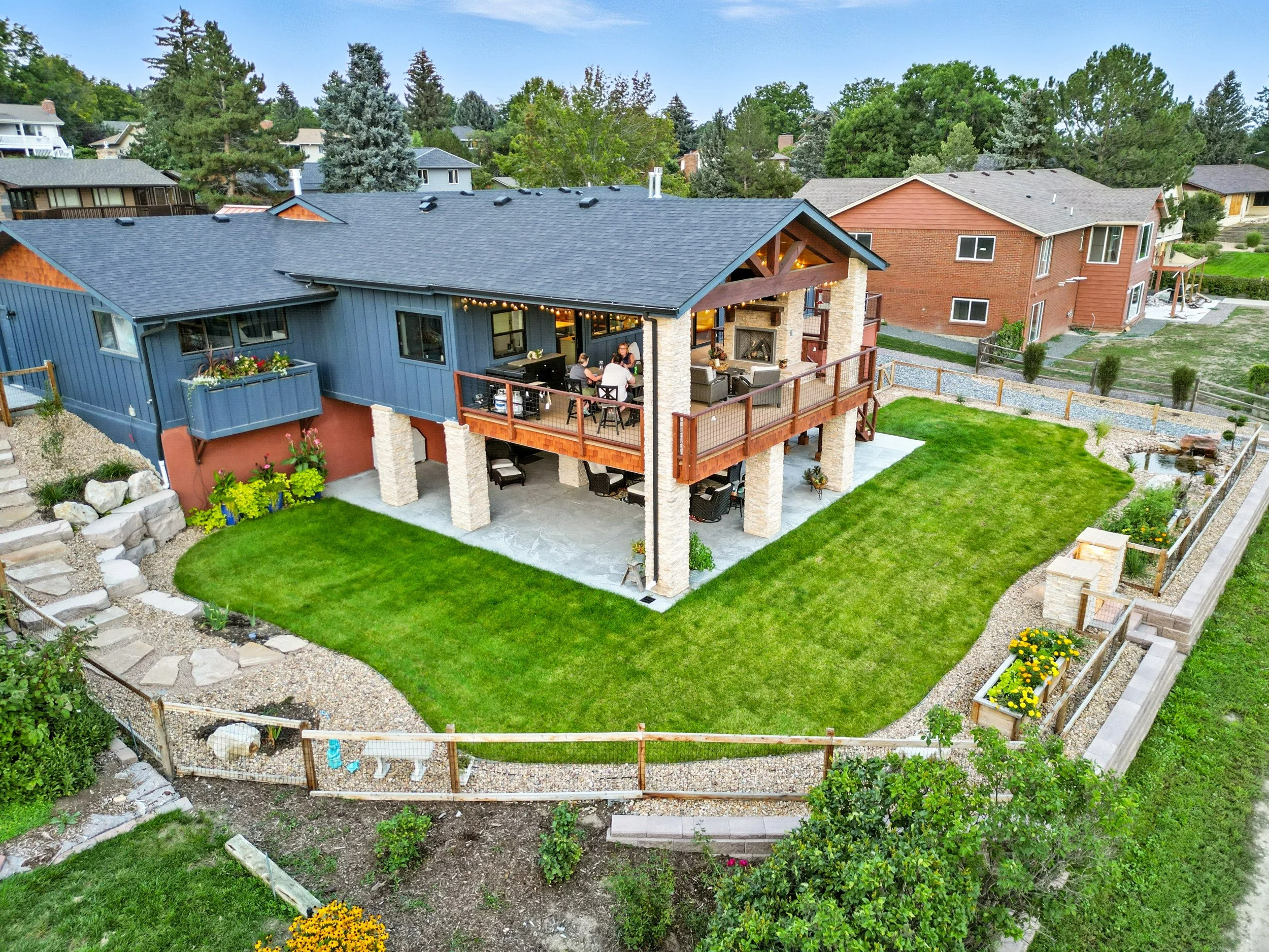 A large backyard with a green lawn, stone path, and fenced garden, adjacent to a blue and brick house with a spacious balcony and an outdoor seating area.