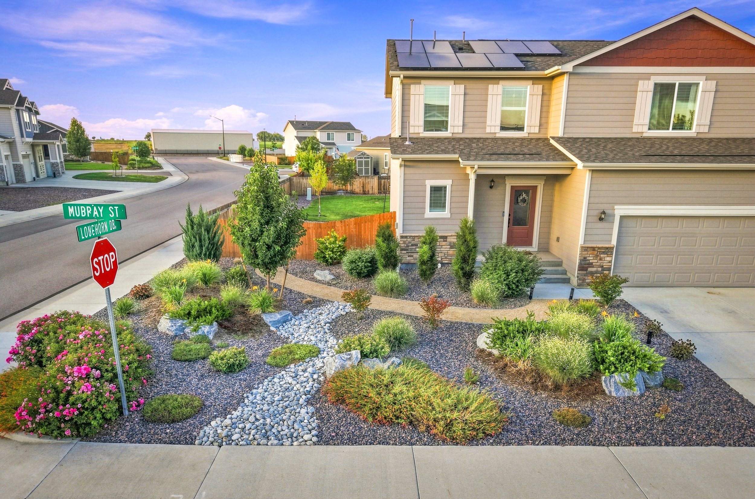 A landscaped front yard with colorful flowers, small trees, and decorative rocks in a suburban neighborhood. A two-story house with solar panels on the roof is visible in the background. A street sign indicates the intersection of MUBPAY ST and LONEH
