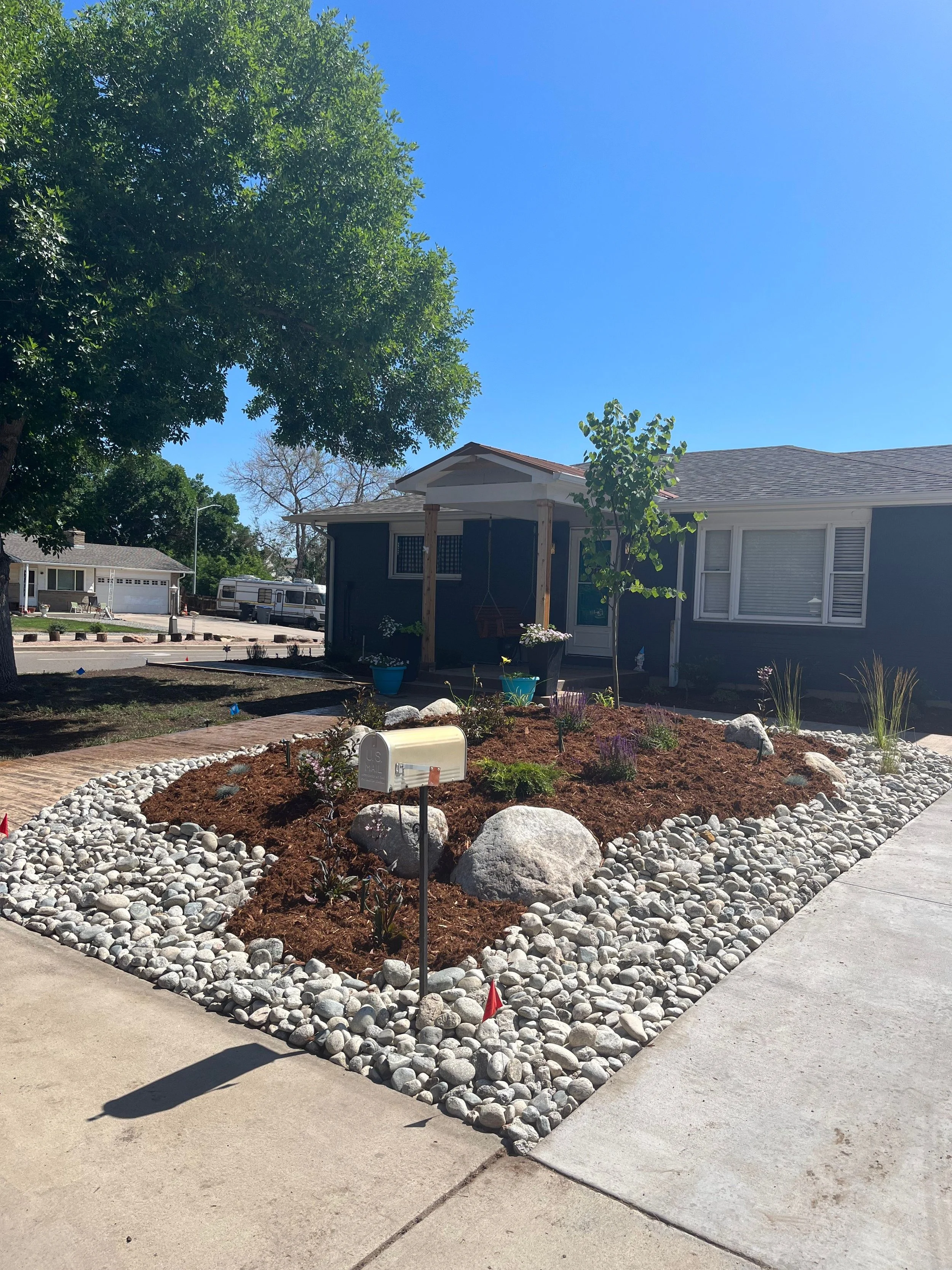 A residential front yard with freshly planted garden beds edged with white rocks, a small tree, and potted plants. A mailbox is near the sidewalk. The house is dark-colored with white trim and a small porch. There are other houses and a parked traile