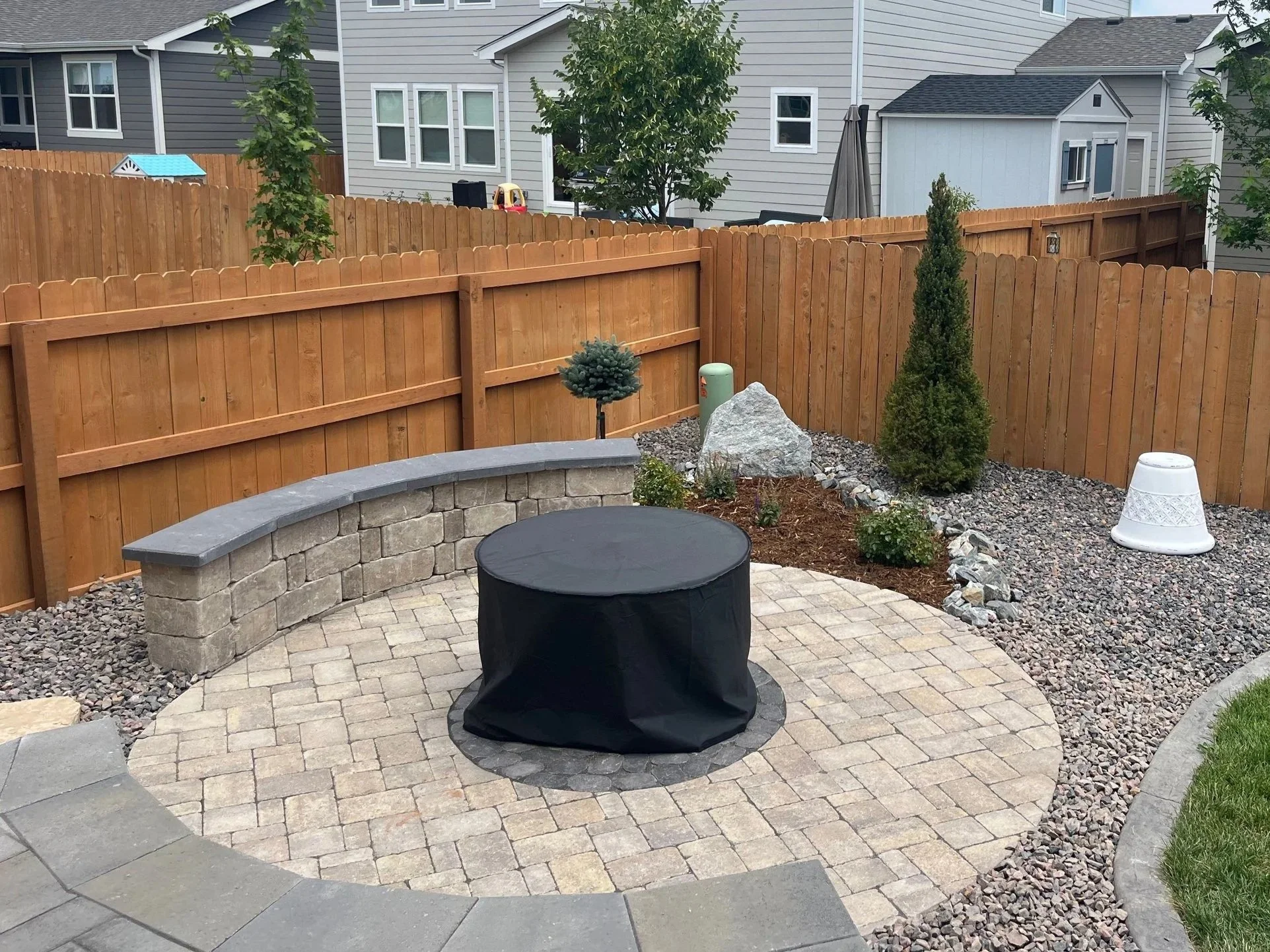 Backyard patio with a stone paver circle, a small black table covered with a black cloth, a low stone wall, and a wooden fence, with trees and house in the background.