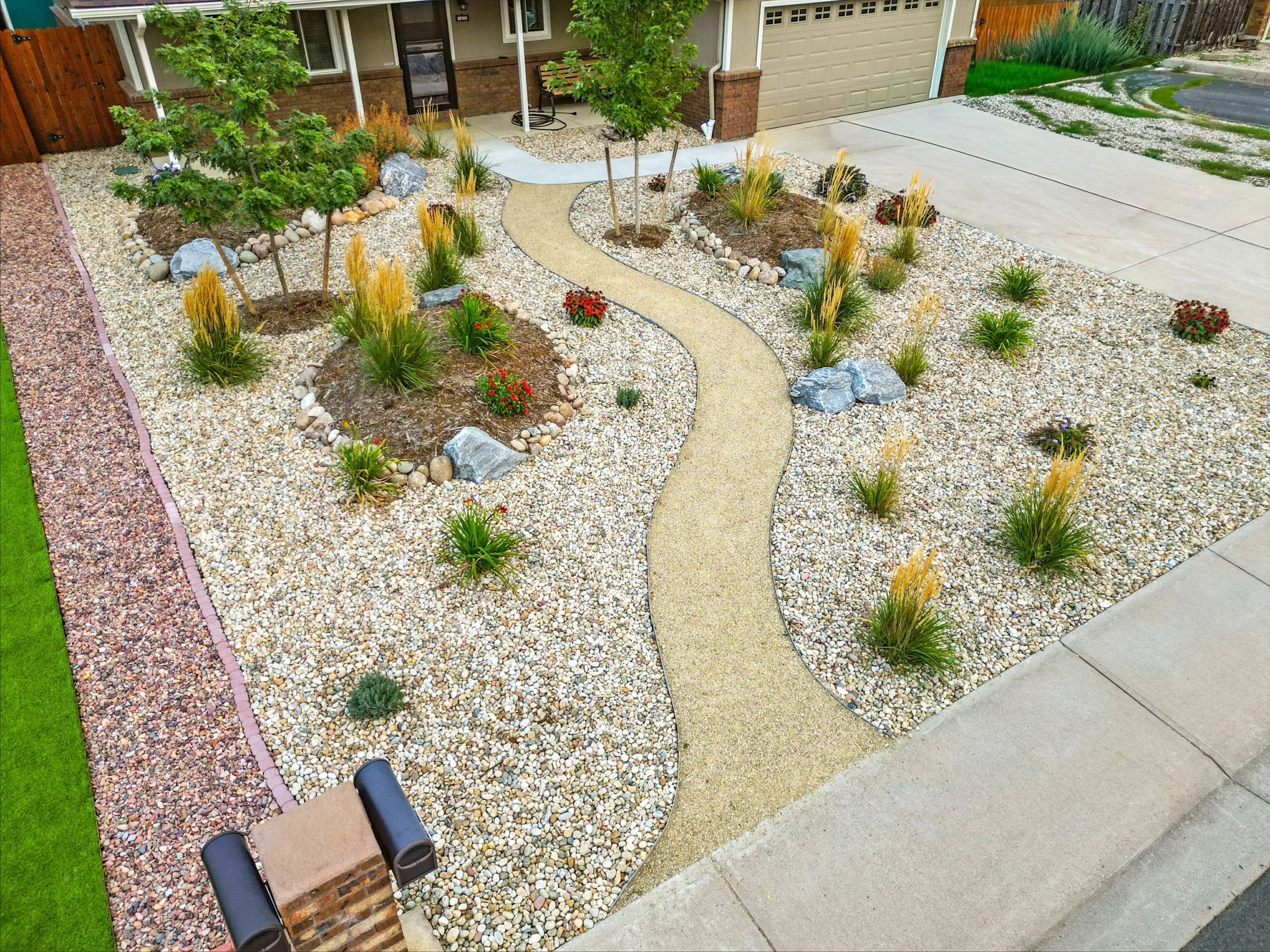 A landscaped front yard with a winding concrete walkway, surrounded by small rocks, grasses, and flowering plants. The yard features two small trees and is bordered by a wooden fence and a house with a garage.