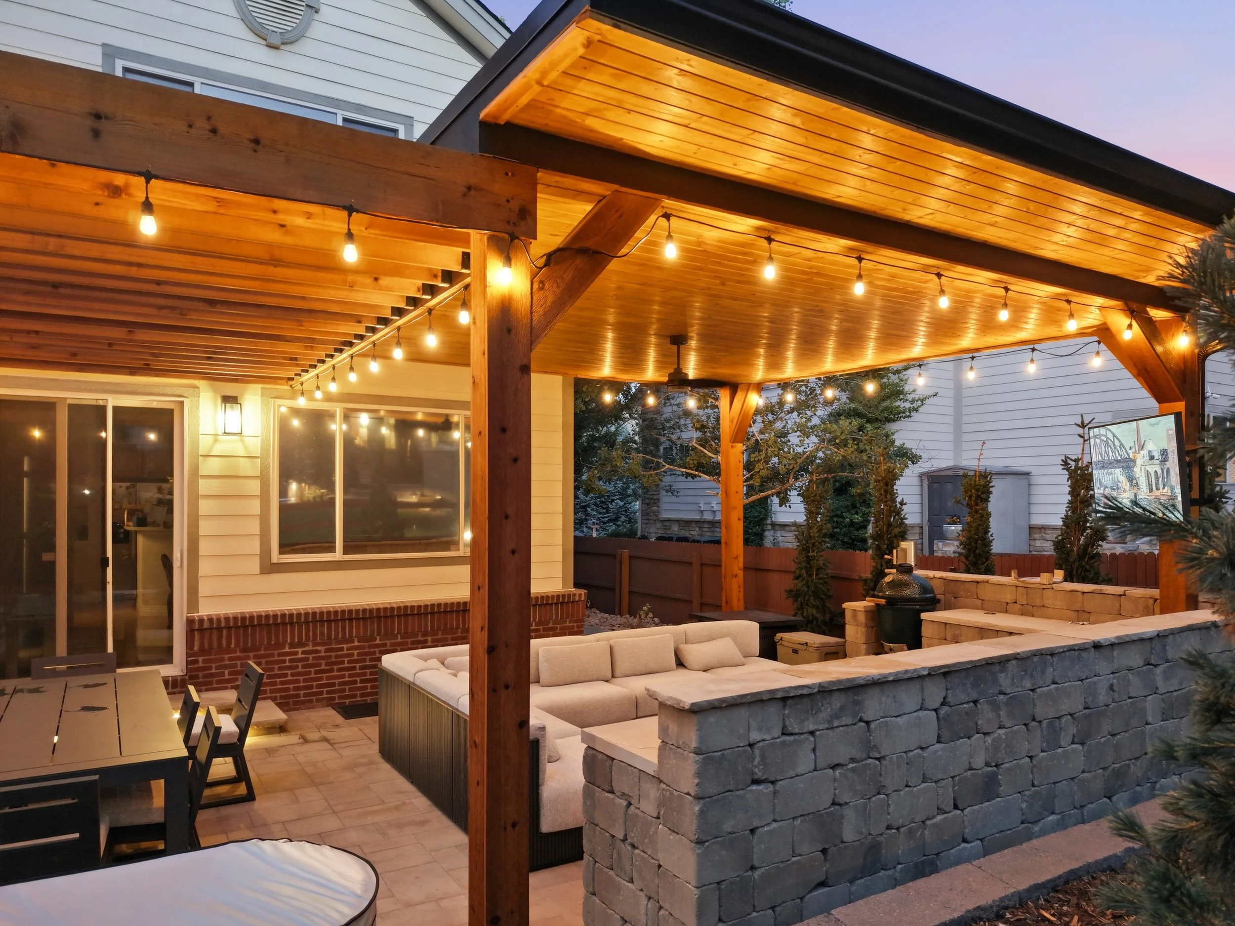 Outdoor patio area with wooden pergola, string lights, a seating area with cream cushions, a dining table with chairs, brick and stone wall features, and a view of neighboring houses.