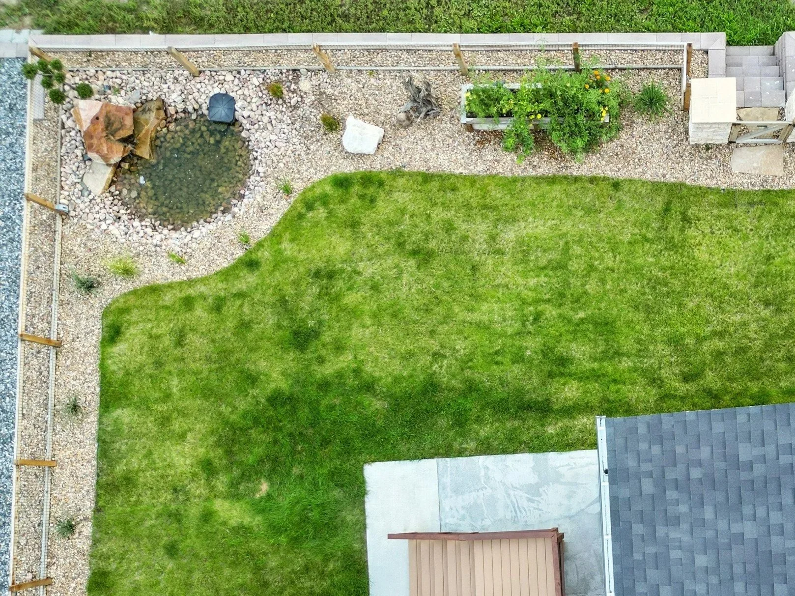An aerial view of a backyard with a small pond surrounded by rocks, a green grassy lawn, a gravel area with plants and garden decorations, and a concrete patio with a wooden step.
