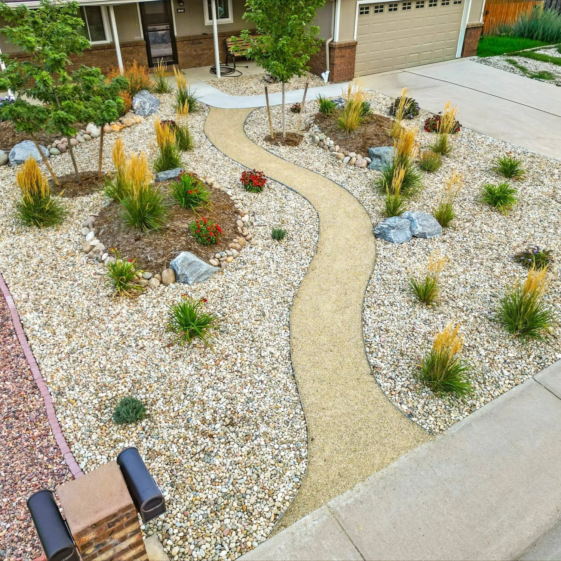 A landscaped front yard with a curved concrete pathway, small trees, drought-tolerant plants, and decorative rocks, adjacent to a house with a garage.