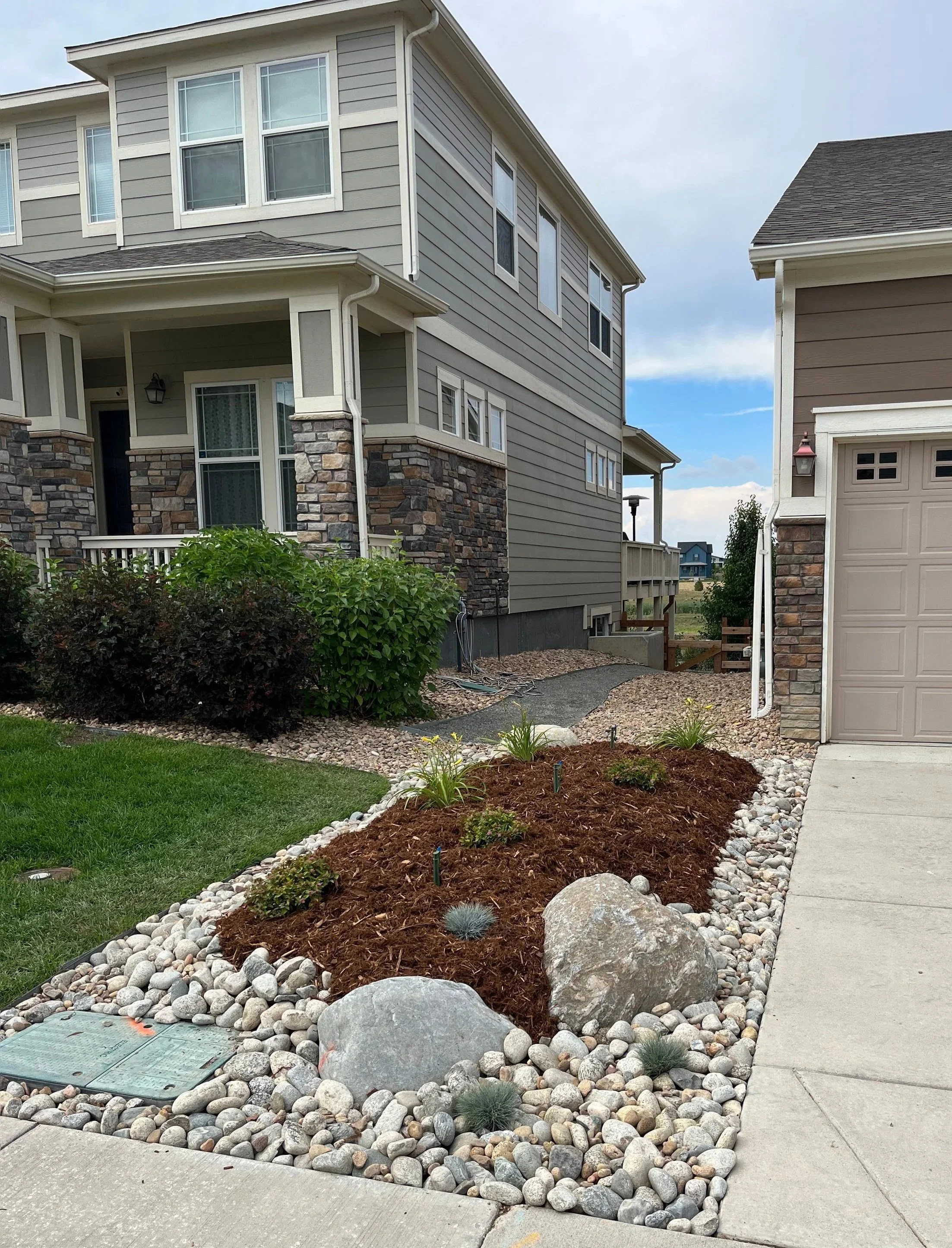 Front yard with a landscaped garden bed containing mulch, rocks, small plants, and large stones, next to a concrete sidewalk and driveway, in front of a modern multi-story house with gray siding, stone accents, and several windows.