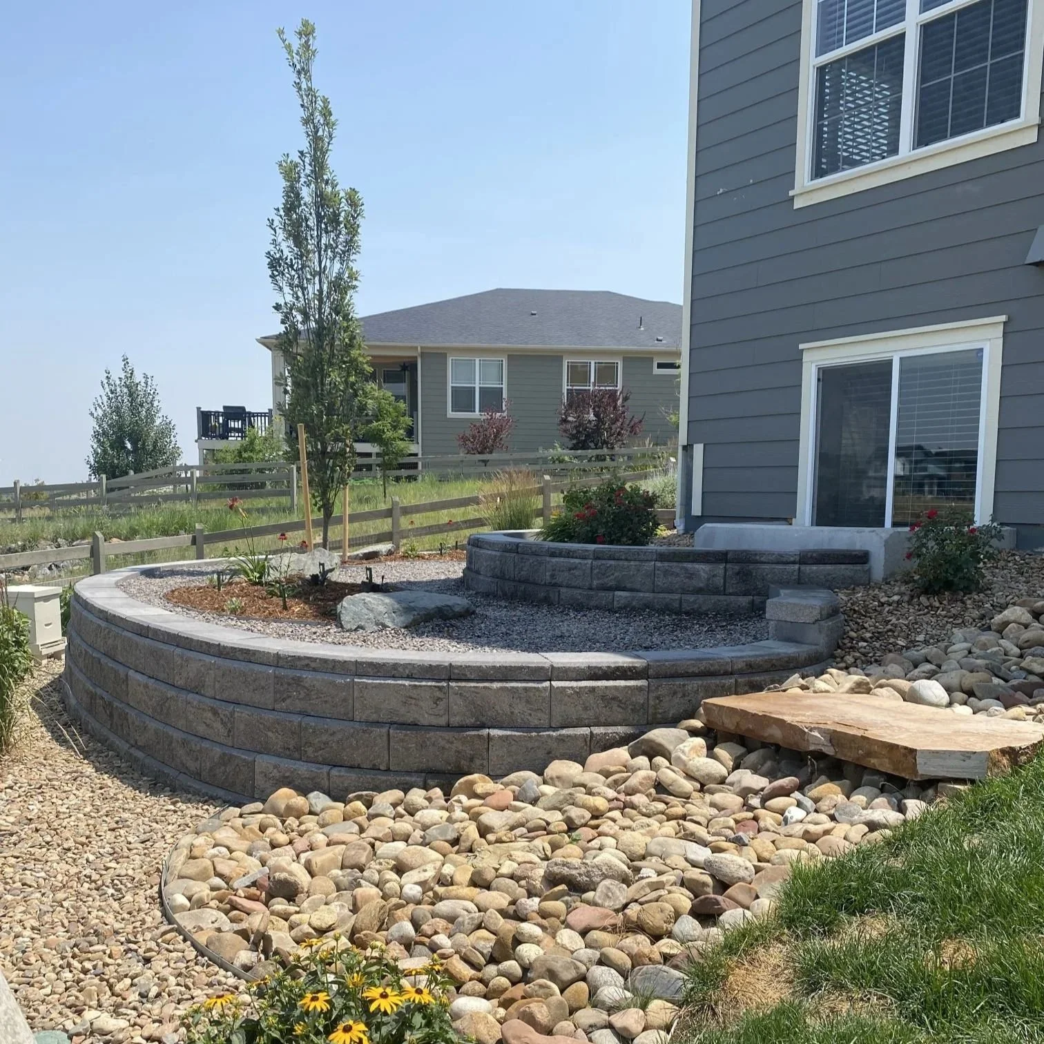 A landscaped backyard garden with layered stone retaining walls, a small tree, plants, and rocks near a gray house with sliding glass doors.