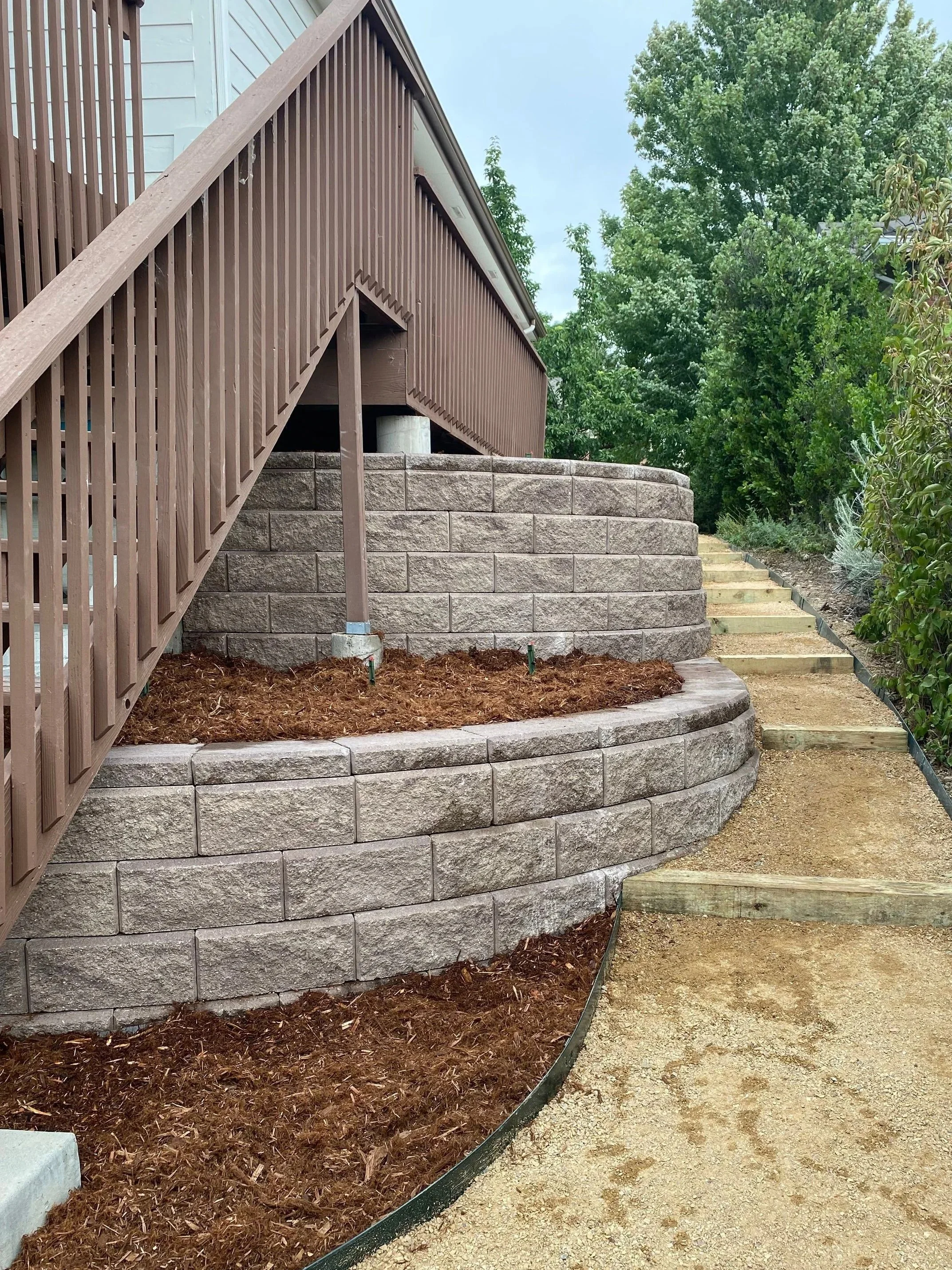 Newly constructed tiered retaining wall made of concrete blocks, along with a staircase with wooden steps, all beside a house with a brown wooden deck and surrounded by green trees.