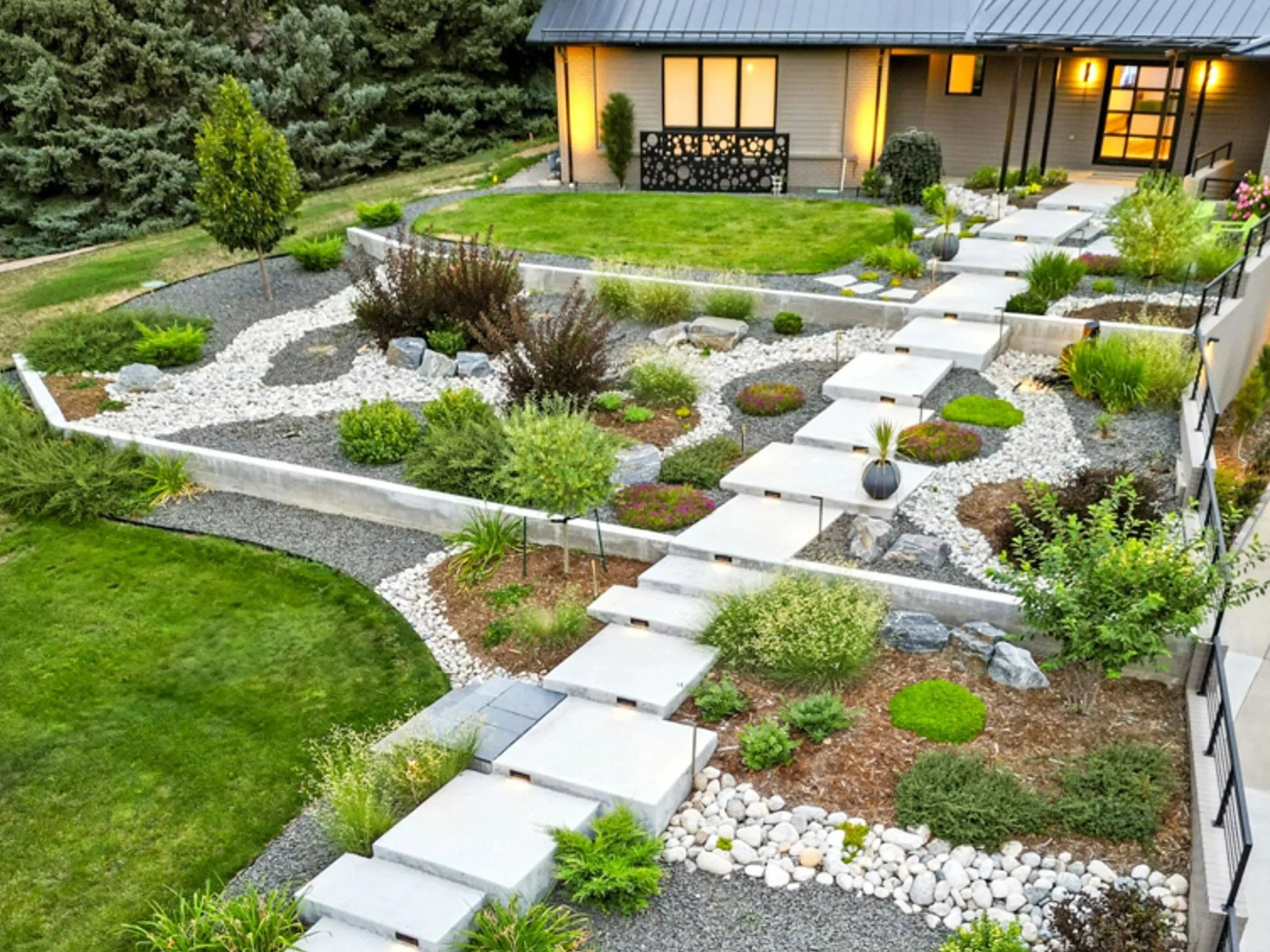 Beautiful backyard garden with a modern pathway, well-manicured lawn, various shrubs, plants, and decorative rocks, adjacent to a house with warm lighting.