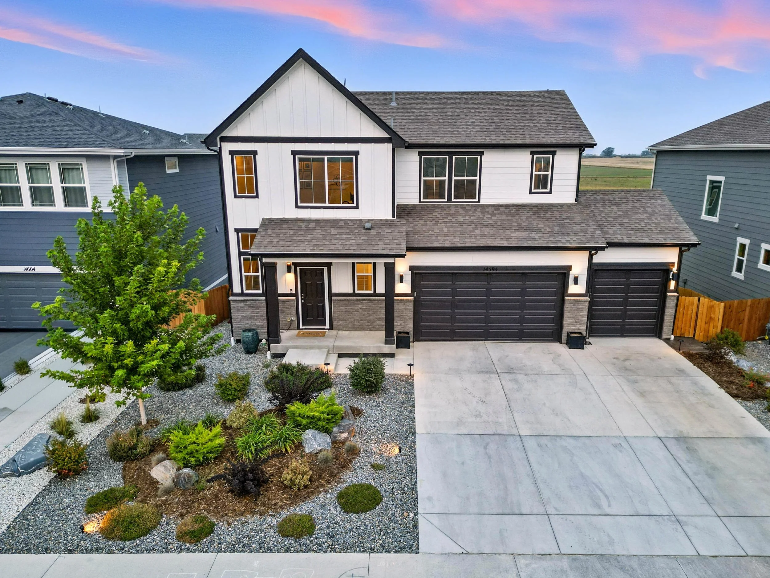 Modern two-story house with black and white exterior, attached three-car garage, landscaped front yard with small tree and rocks, neighboring houses, and an open field in the background during sunset.