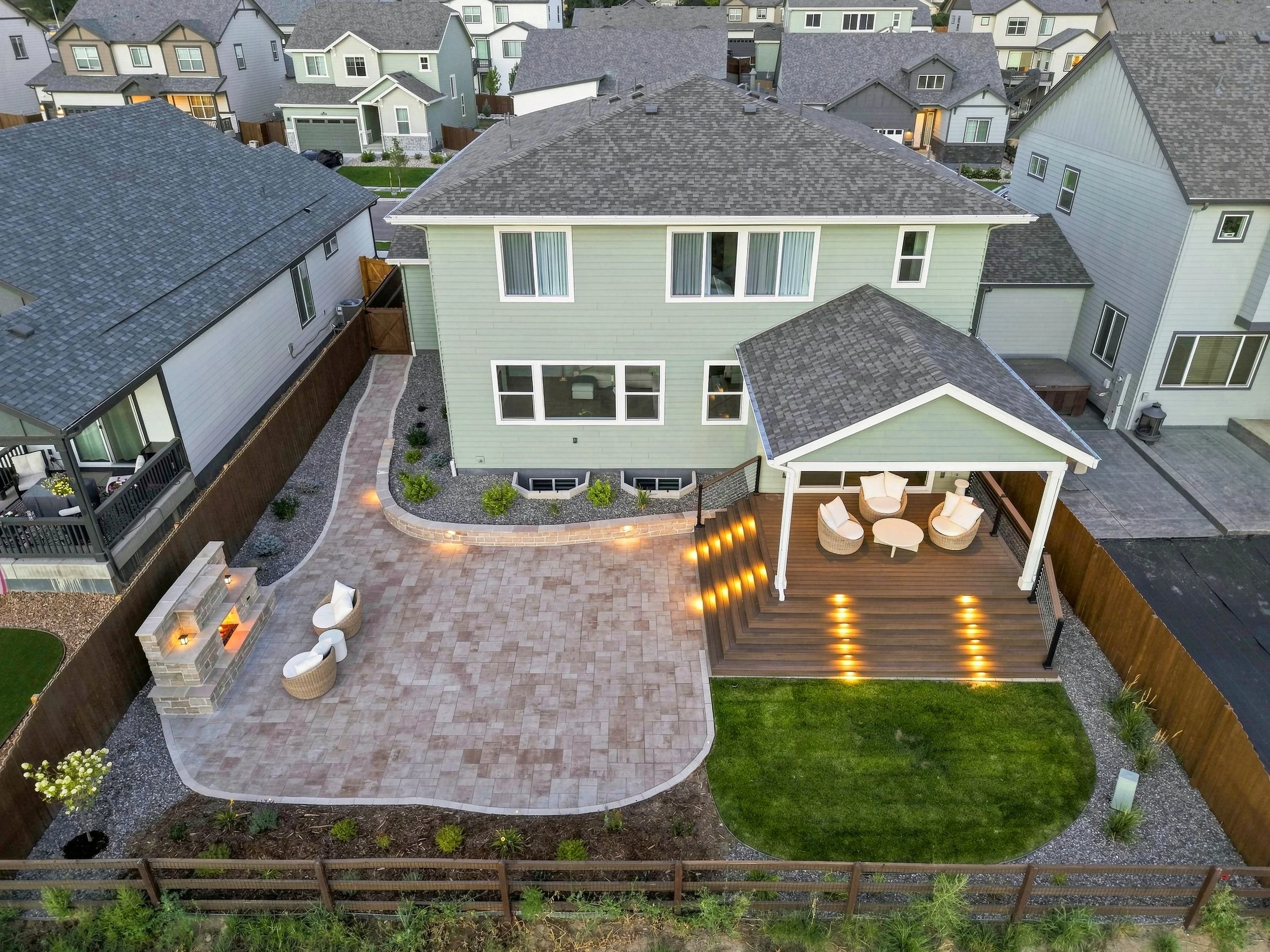 Aerial view of a backyard with a paved patio area, outdoor seating, newly planted lawn, and neighboring houses with similar roofing, fences, and some outdoor furniture and decorative lights.