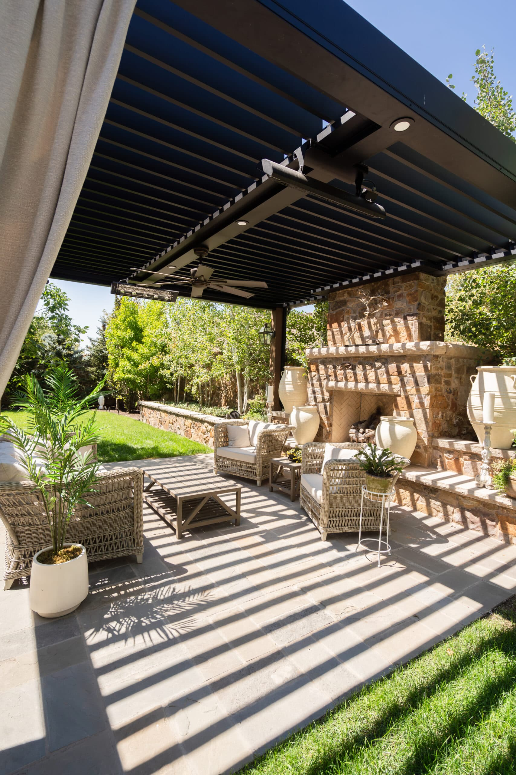 A covered outdoor patio with wicker furniture, potted plants, a stone fireplace, and a ceiling fan, with a garden and trees in the background and shadows cast on the patio floor.