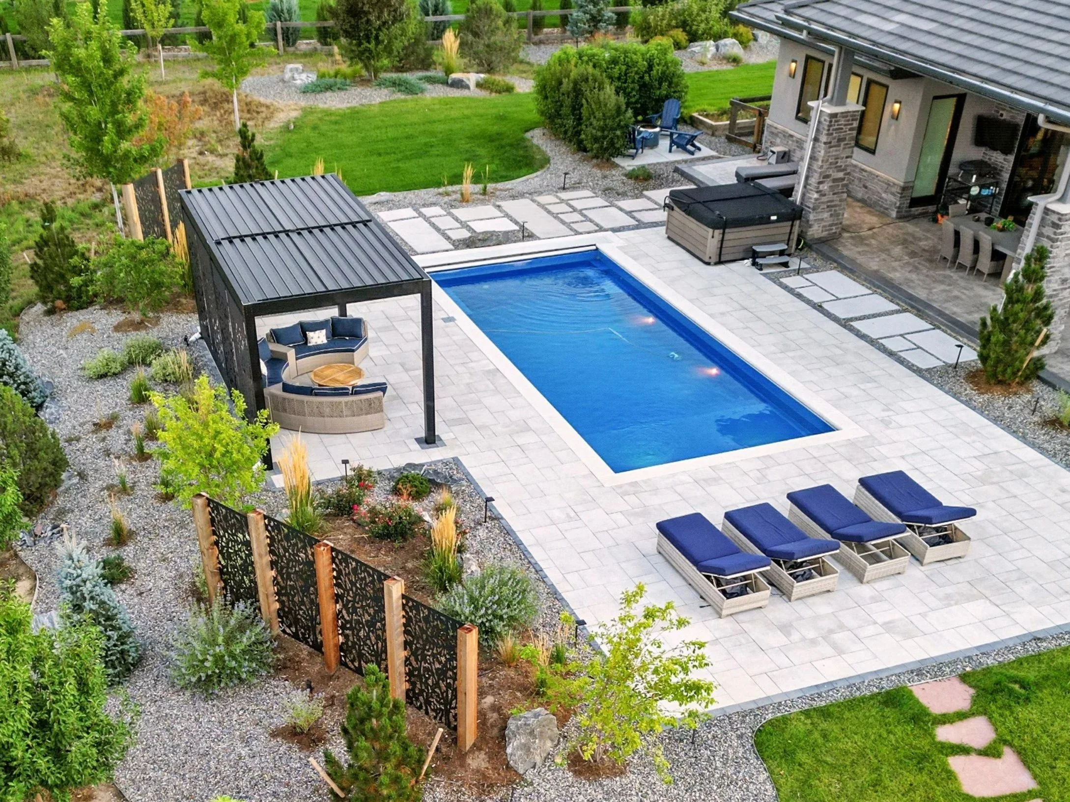 Backyard with rectangular swimming pool, four lounge chairs with blue cushions, a shaded seating area with a curved sofa and a table, hot tub, pathway, outdoor dining, garden with trees and shrubs, and a house with brick and siding exterior.