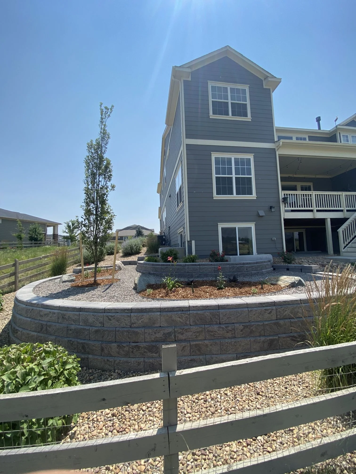 A multi-story gray house with white trim, a second-floor balcony, and landscaped yard with trees, flowers, rocks, and a fence under a clear blue sky.