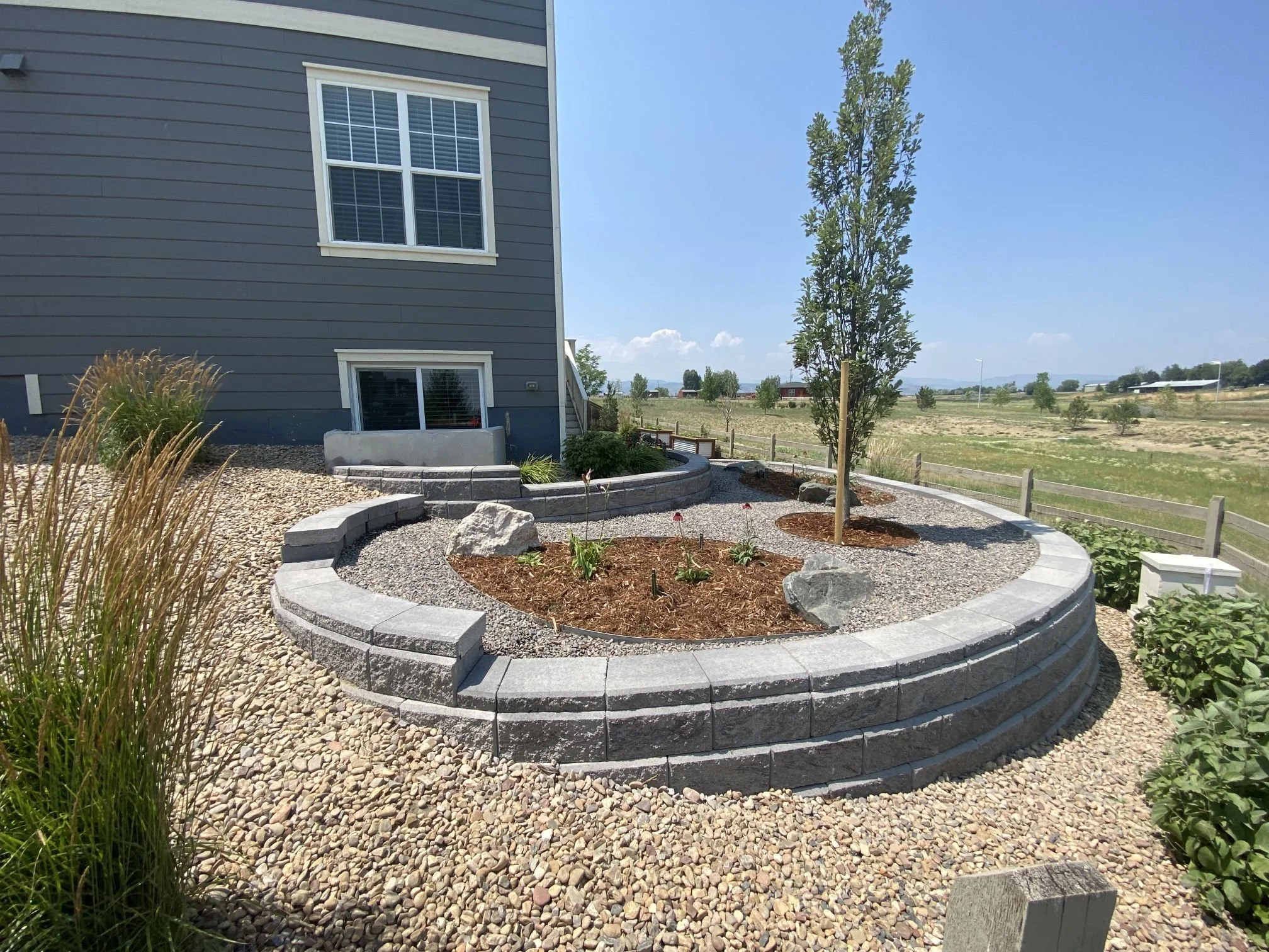 A landscaped yard with a curved retaining wall made of gray bricks, containing a small tree, flowers, and mulch, with a house in the background and an open field beyond.