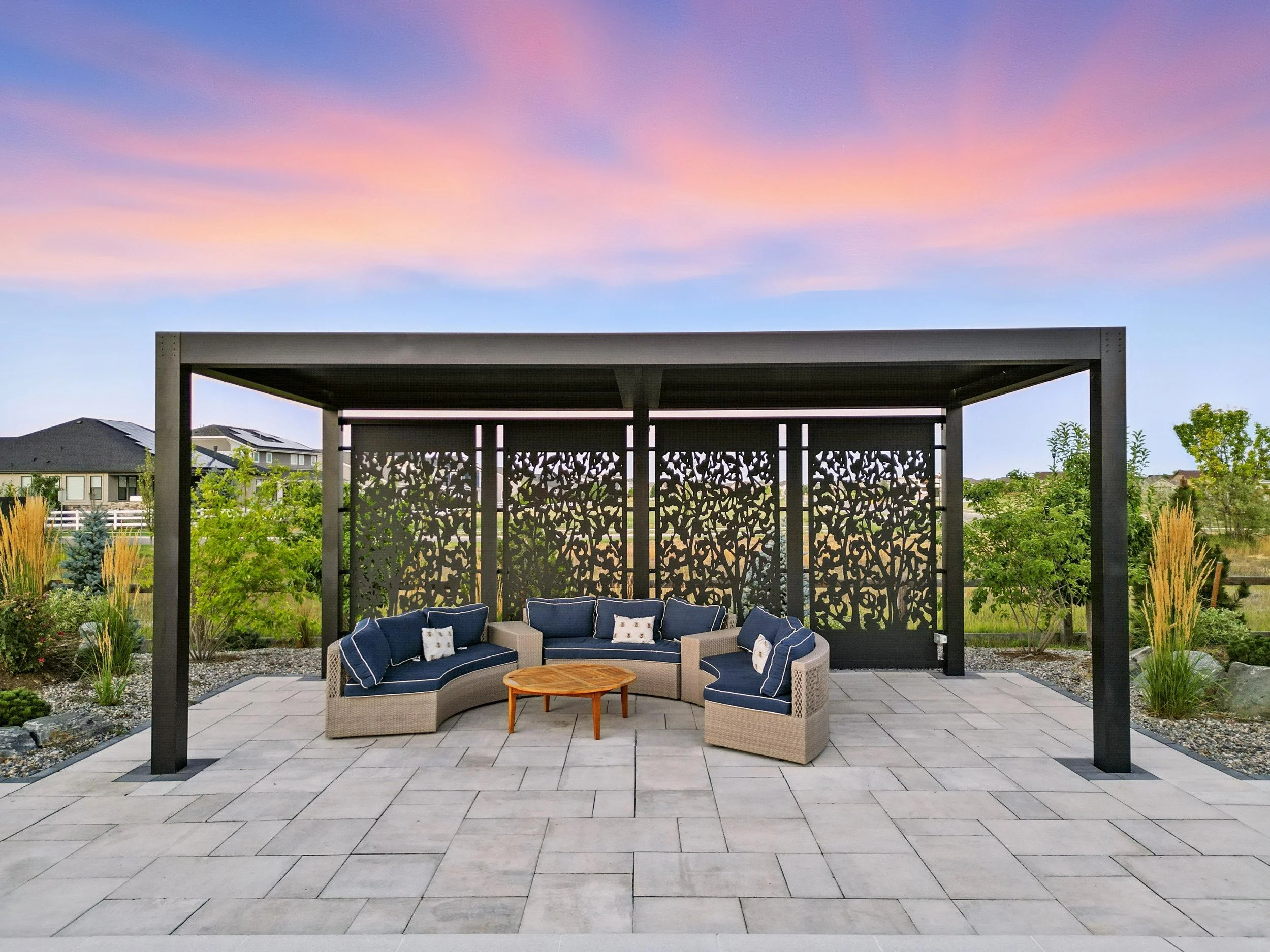 outdoor seating area with a sectional sofa and coffee table under a black pergola, with a decorative black wall and a sunset sky in the background
