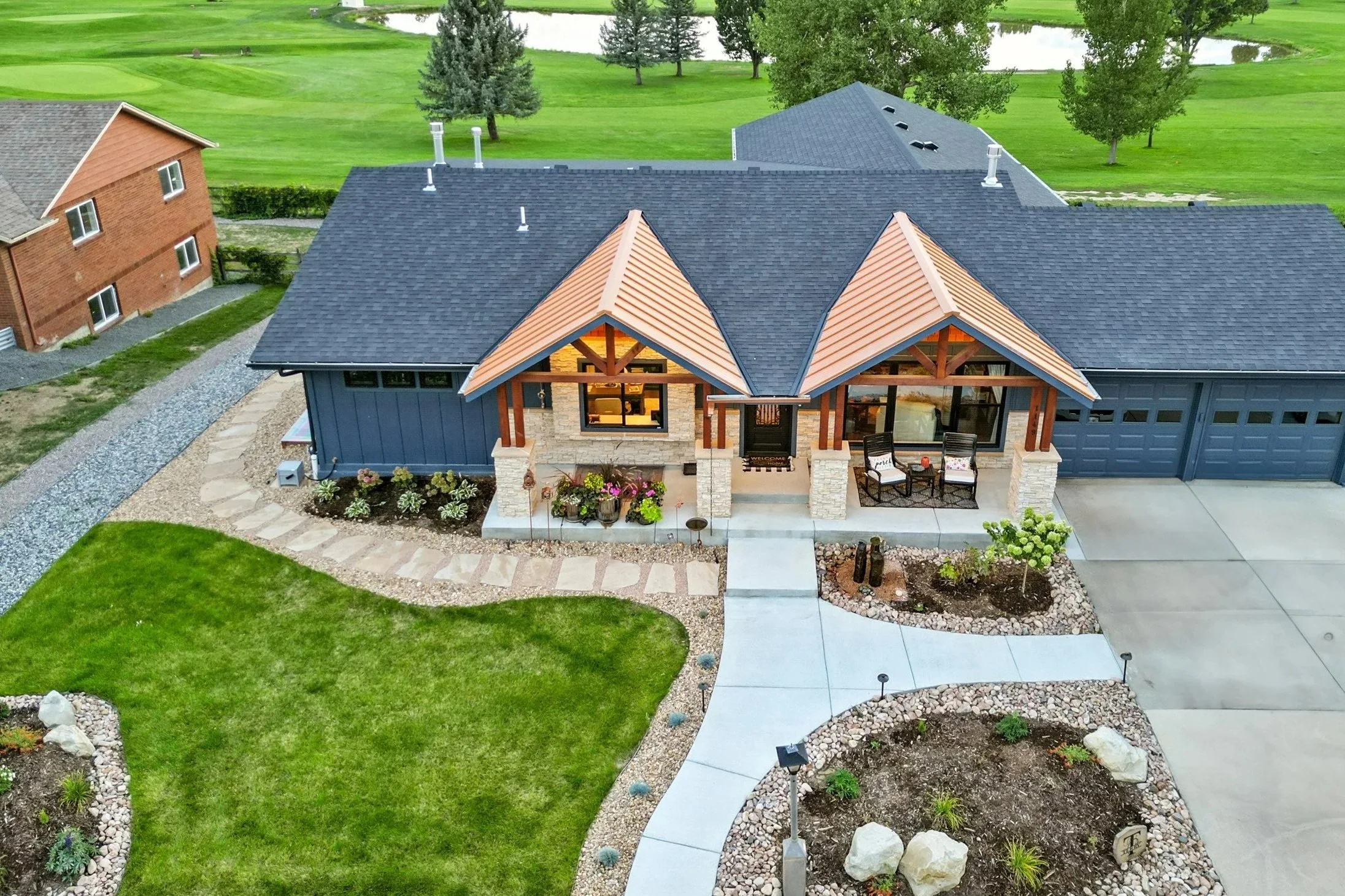 Aerial view of a modern house with a landscaped front yard, a curved walkway, and a large driveway. The house features a dark roof, wooden accents, and stone pillars on the porch. Surrounding the house are green lawns, trees, and neighboring homes.