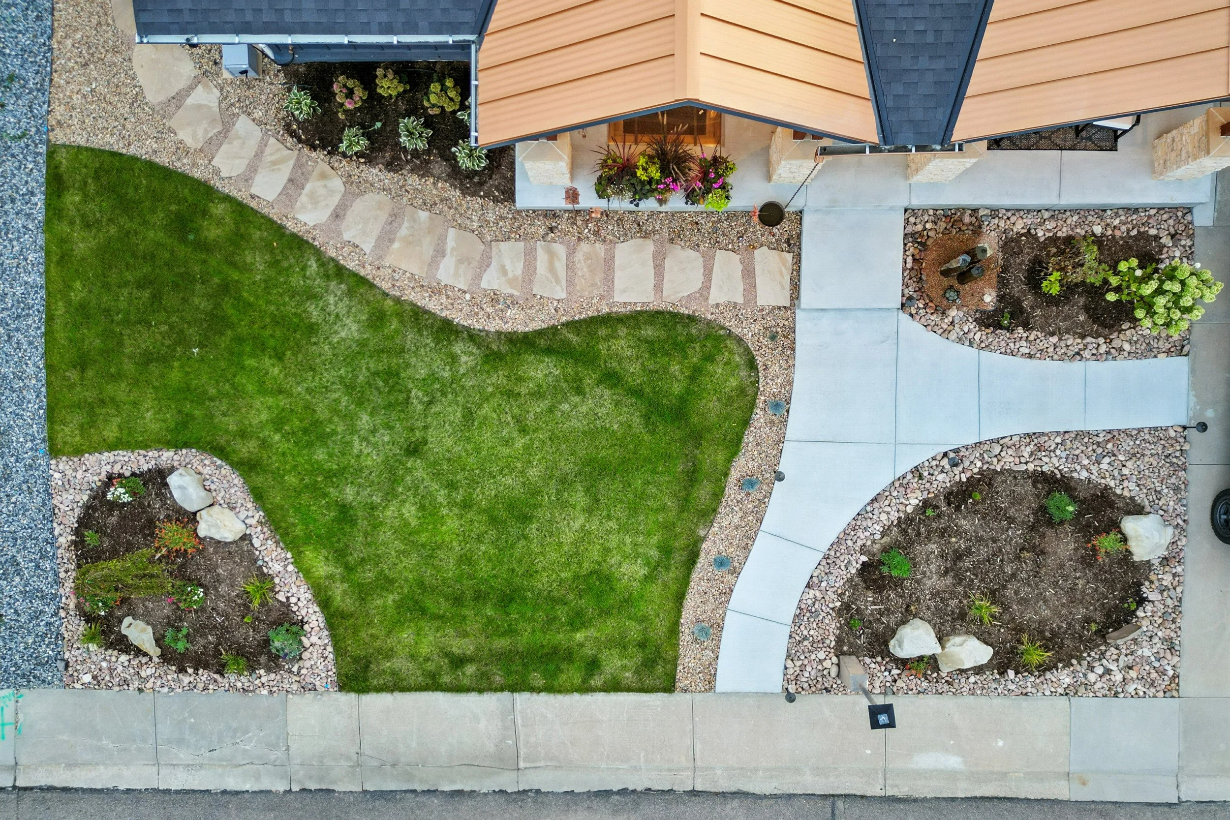 Aerial view of a front yard with a grassy area, stone walkway, flower beds with rocks and plants, concrete pathway, and parts of the house with orange siding and flower pots.