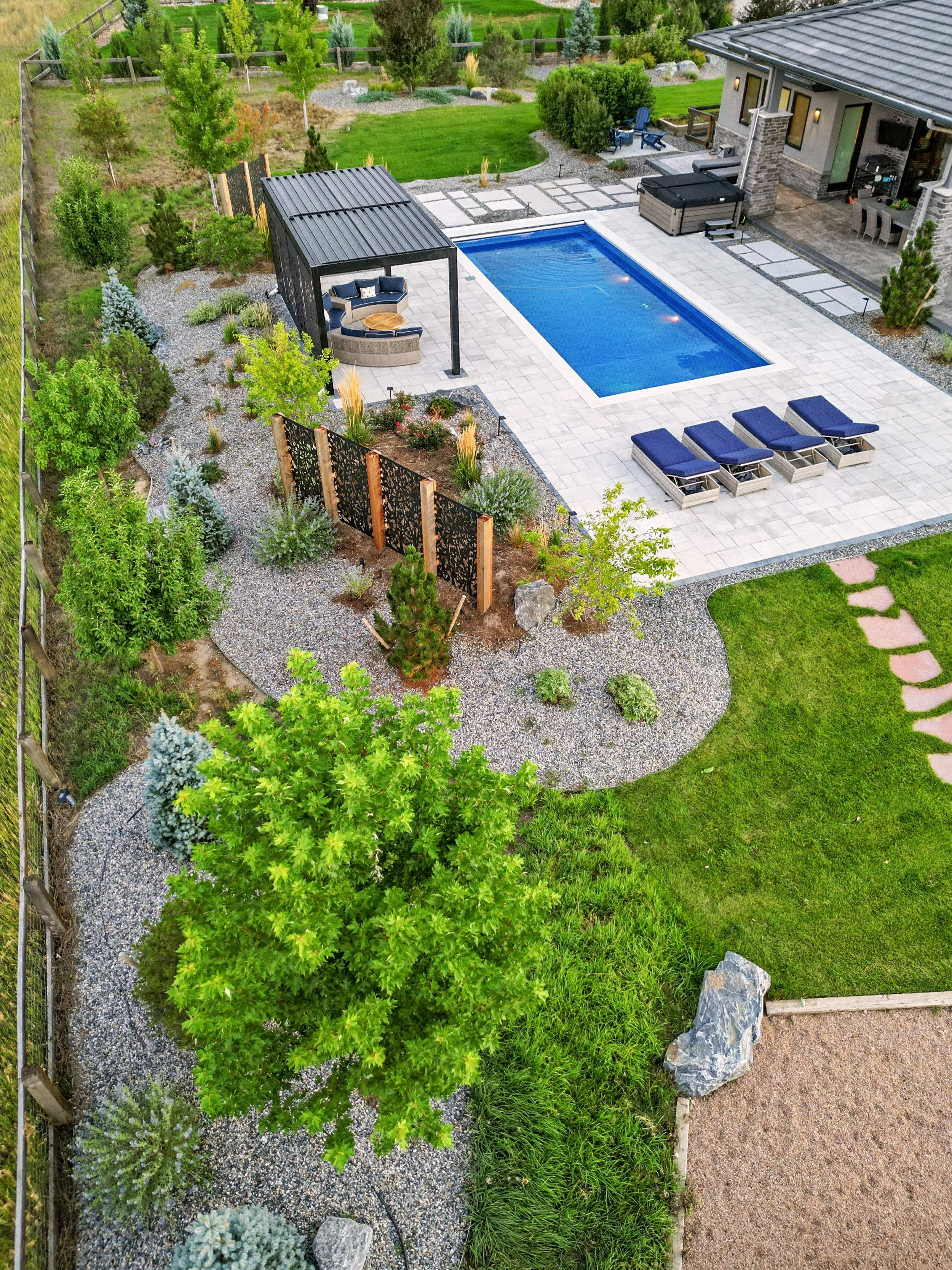 A backyard with a swimming pool, poolside lounge chairs, a pergola with seating, and surrounding landscaped greenery including trees, bushes, and flower beds.