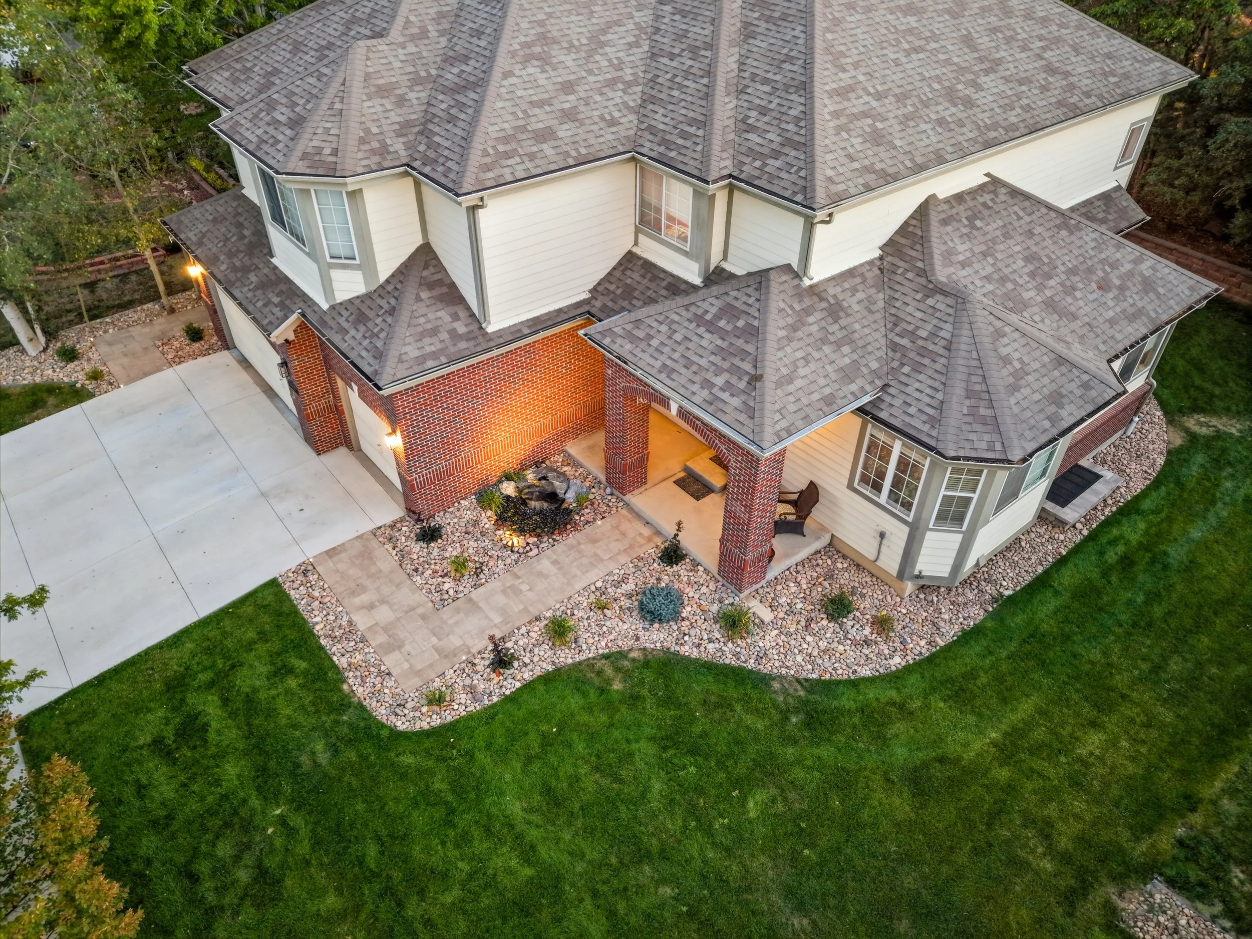 An aerial view of a two-story house with a multi-gabled roof, white and brick exterior walls, large windows, and a surrounding landscaped yard with trees, a concrete driveway, and decorative rocks.