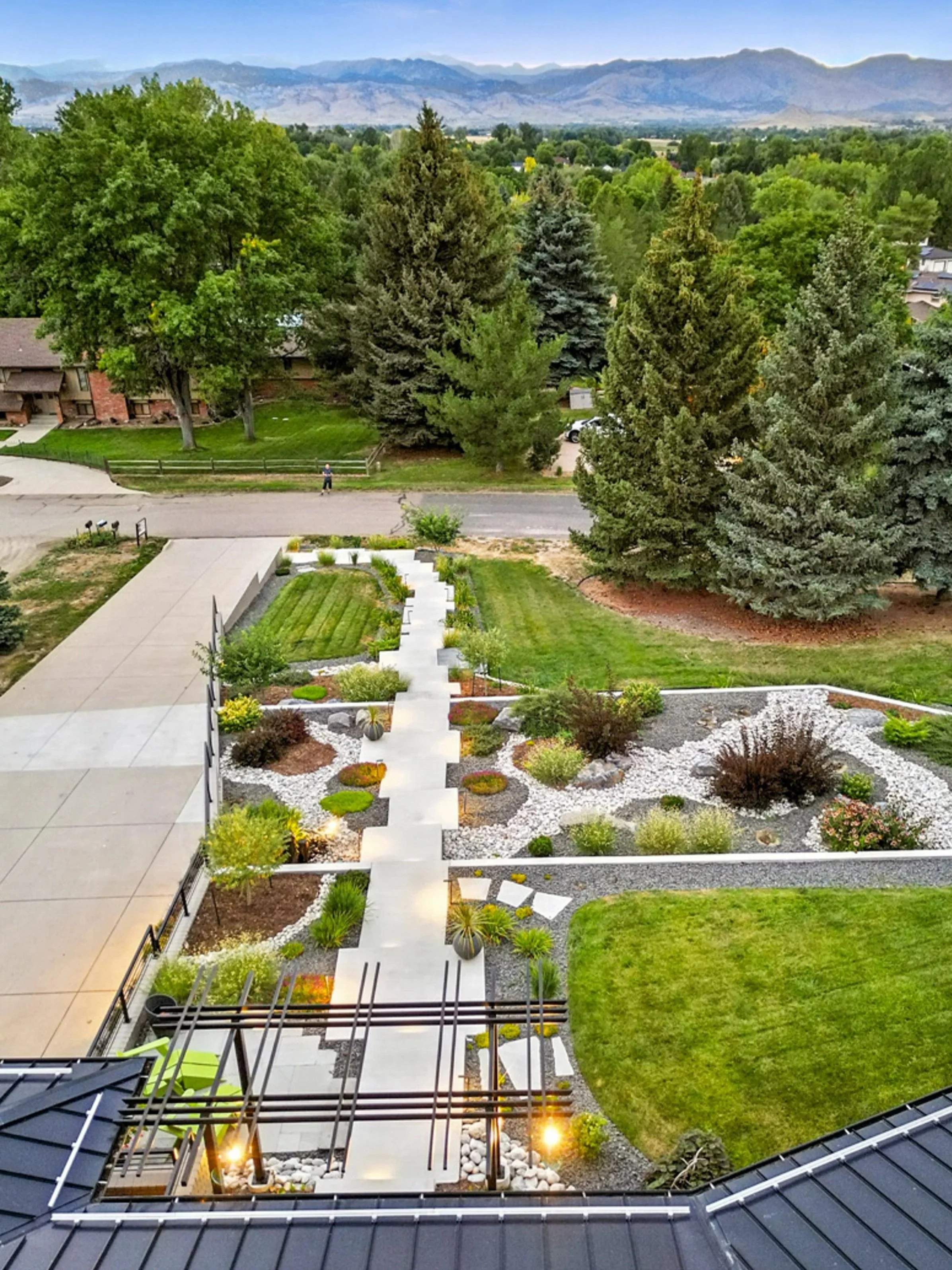 A landscaped backyard with a pathway leading to a lawn, various bushes, succulents, trees, and decorative rocks, with large trees and mountains in the background.