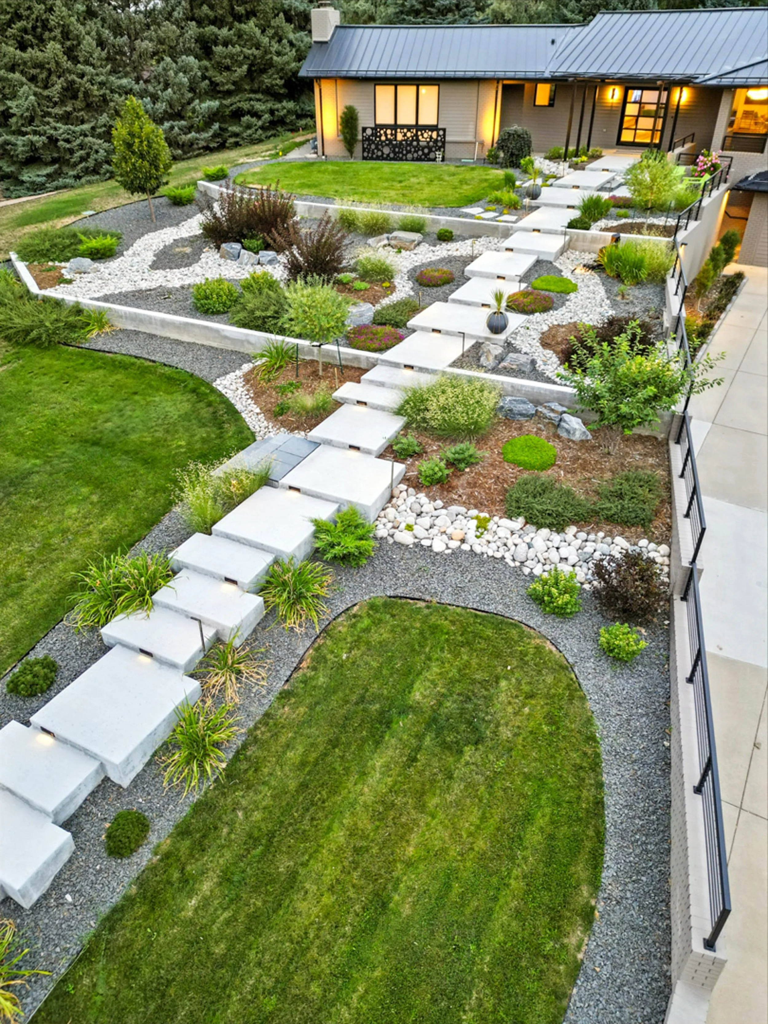 A landscaped backyard with a modern pathway, grass lawn, decorative plants, and trees, adjacent to a house with lit windows.