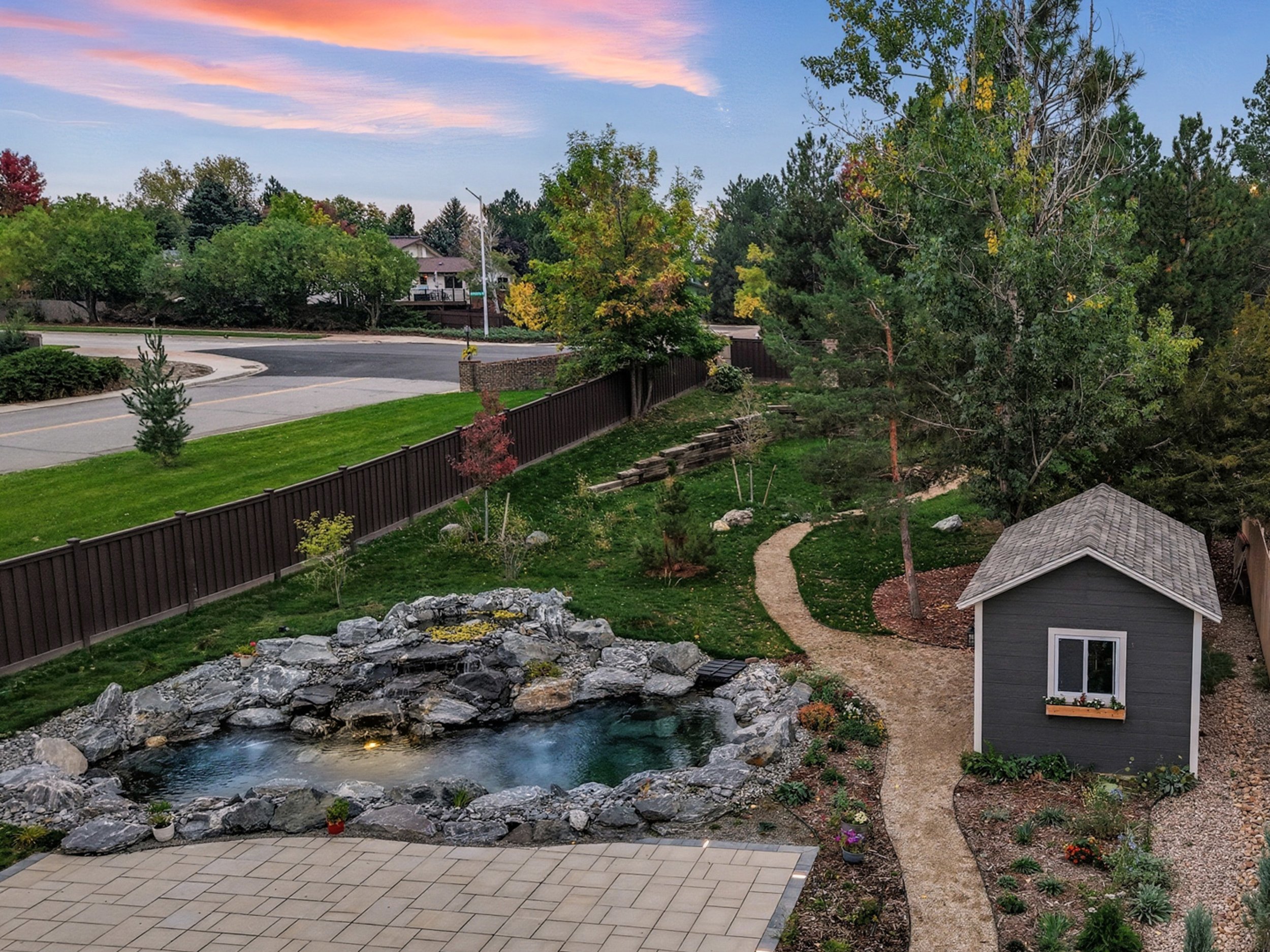 A backyard with a small pond surrounded by rocks, a garden shed, a winding gravel path, and various trees and plants, with a street and houses visible in the background.