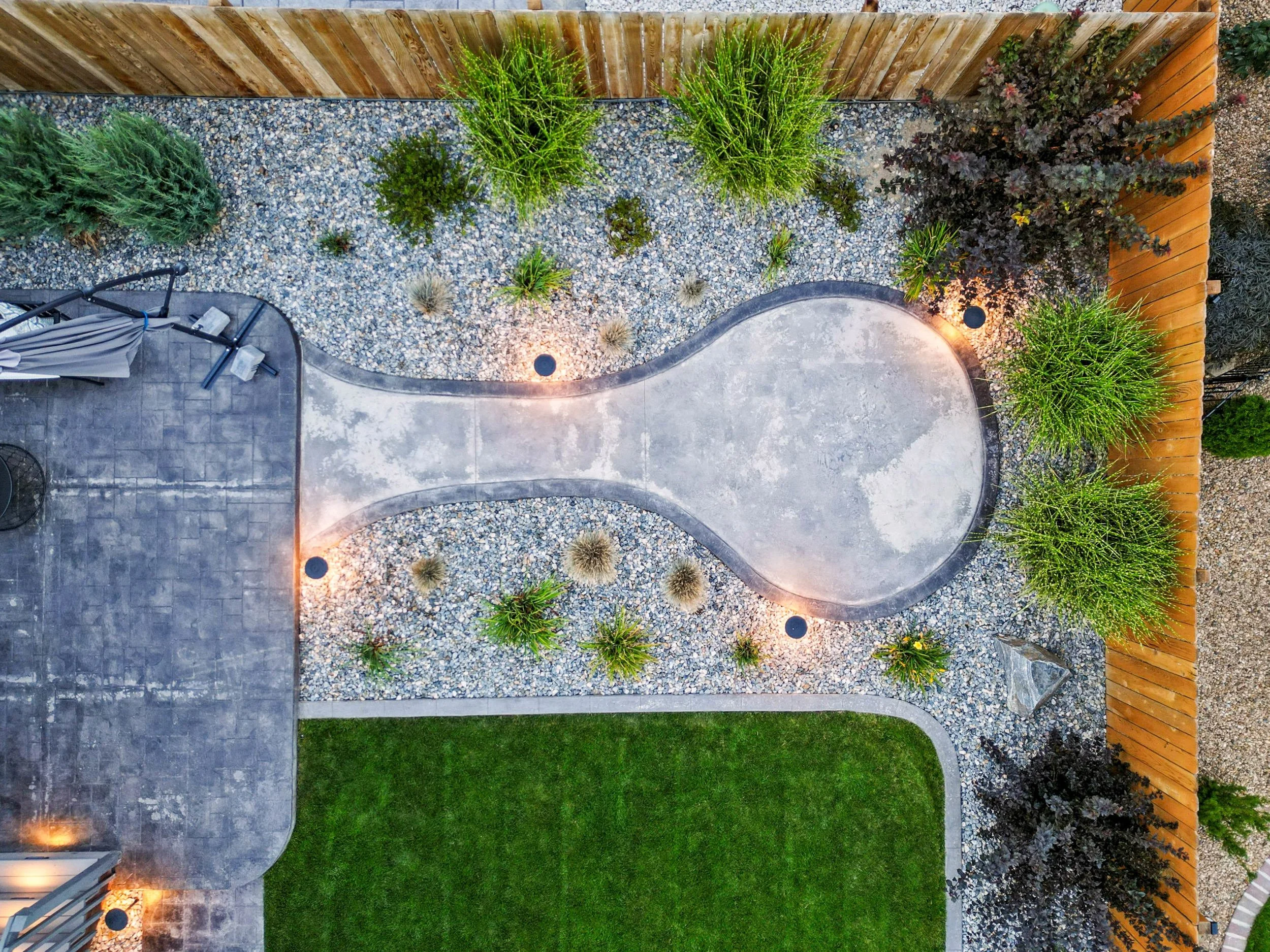 Aerial view of a backyard with a curved concrete walkway, gravel area with various plants, a small grassy patch, a wood fence, and outdoor furniture with umbrellas.