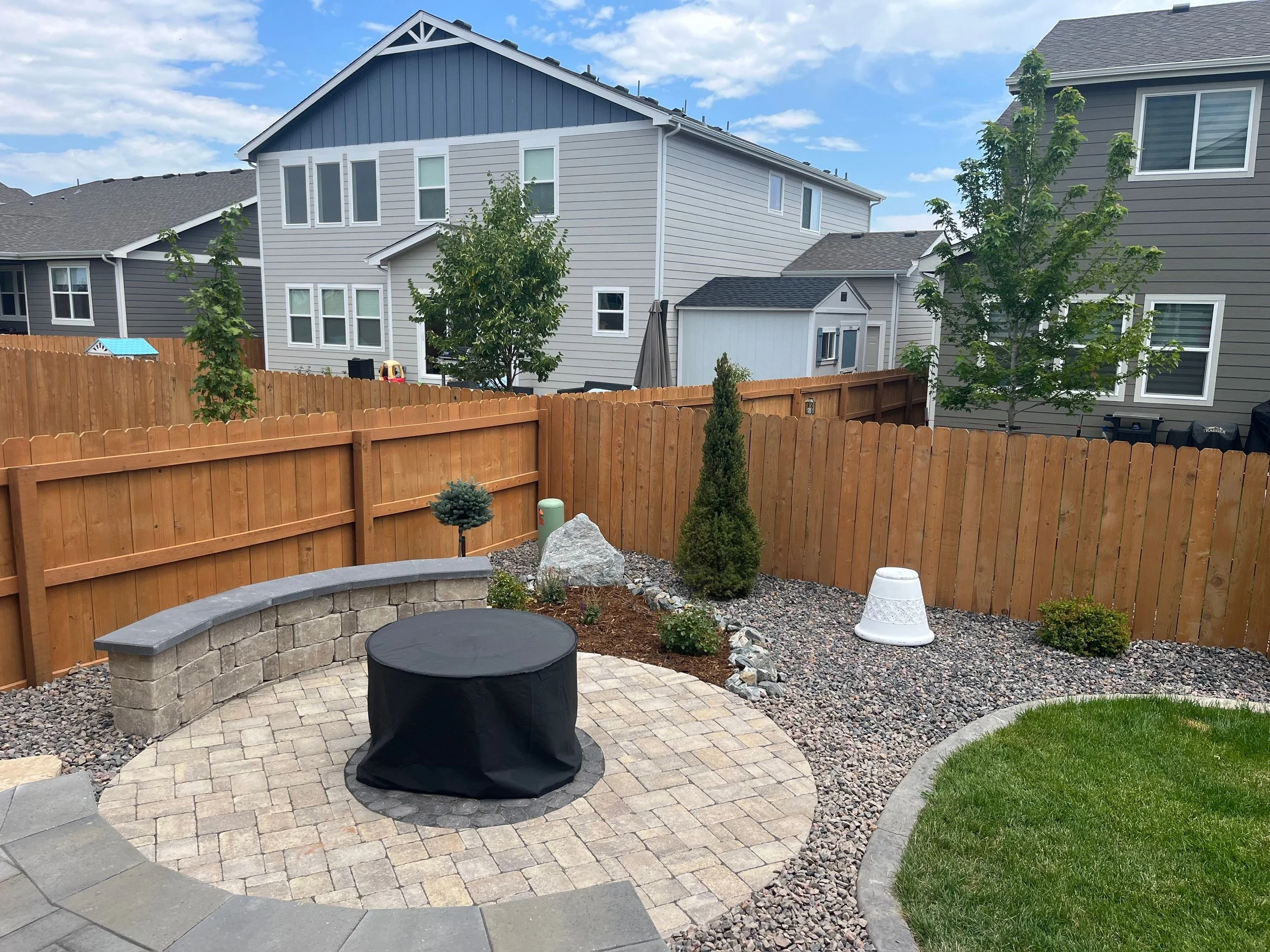 Backyard with a brick patio, a covered table, a curved brick wall, a wooden fence, small trees, shrubs, rocks, and a grassy area.