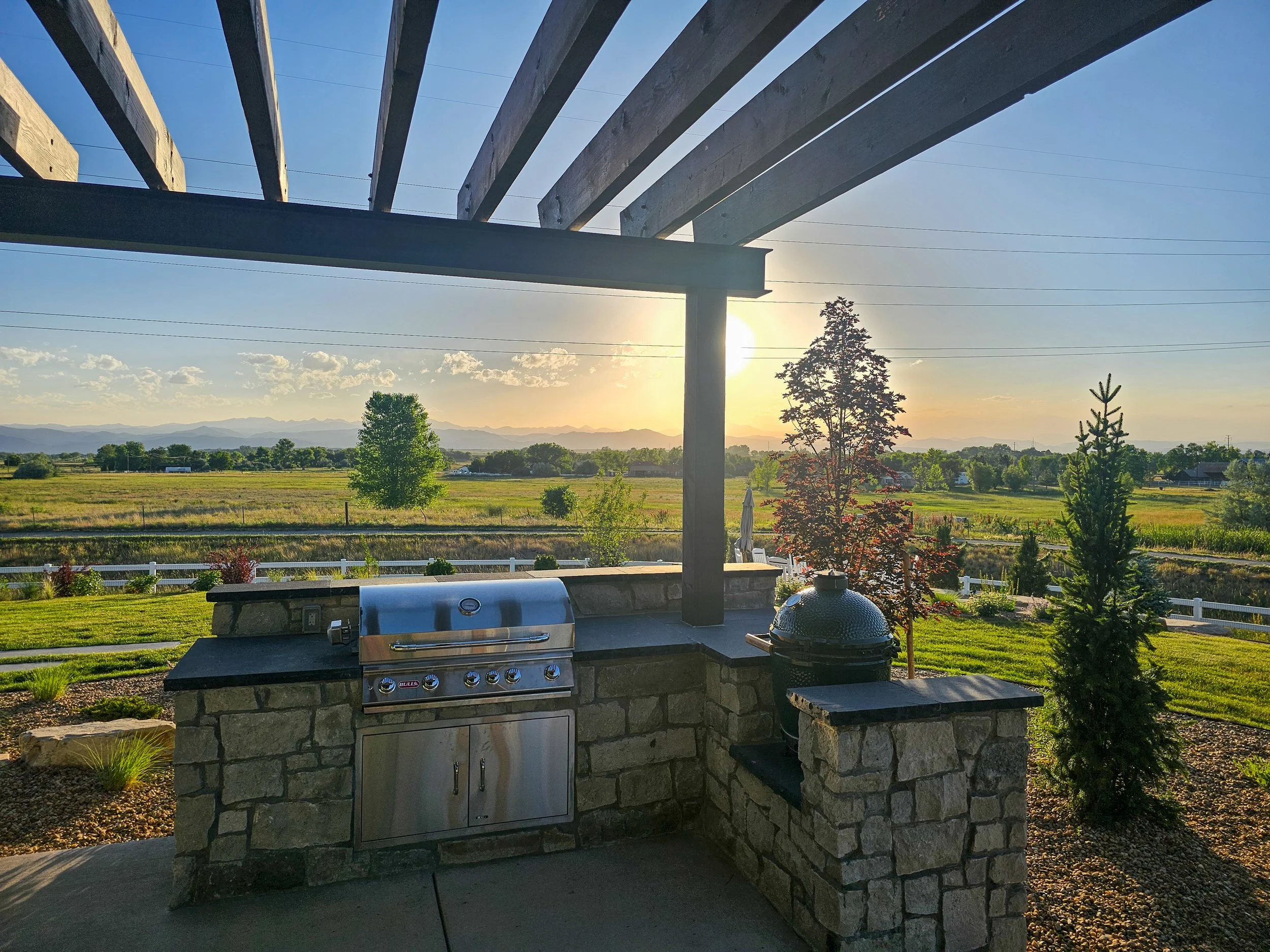 An outdoor kitchen area with a built-in stainless steel grill on a stone counter, a rounded charcoal grill, and a wooden bar or pergola overhead. The background shows open fields, trees, and a mountain range under a sunny sky.