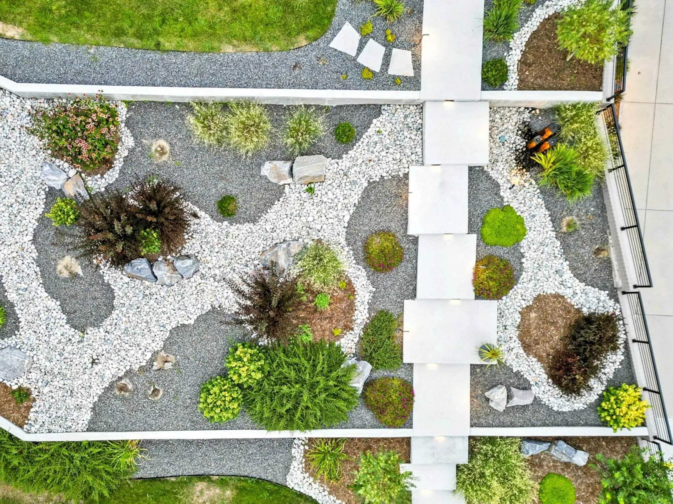 An aerial view of a landscaped garden with winding gravel pathways, various trees and shrubs, white rocks, and concrete walkways, enclosed by a white fence.