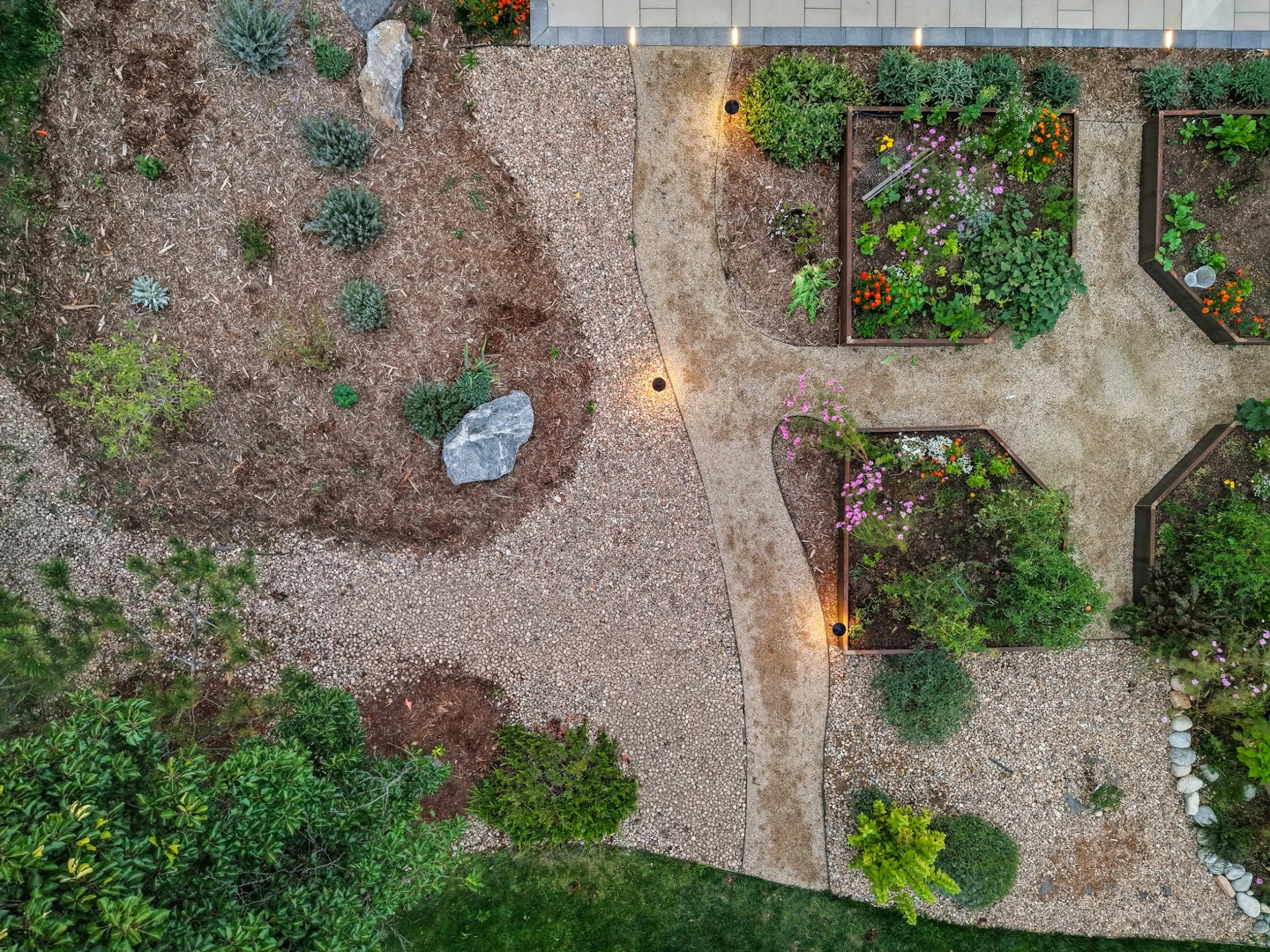 An aerial view of a garden with flower beds containing various colorful flowers, pathways, and landscape elements, including rocks and gravel, illuminated by landscape lighting.